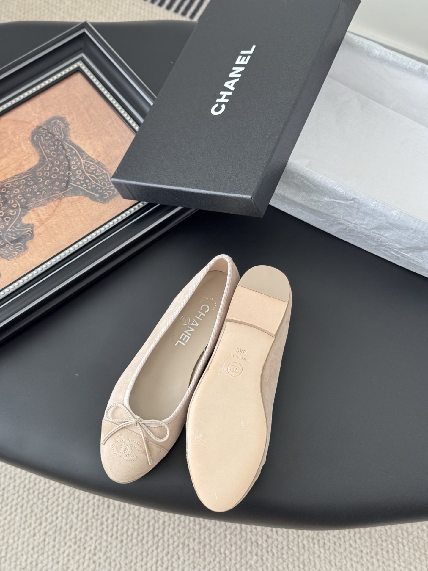 Pair of beige Chanel suede ballet flats with embossed CC logo and bow detail next to a black Chanel shoe box. - Hình 4