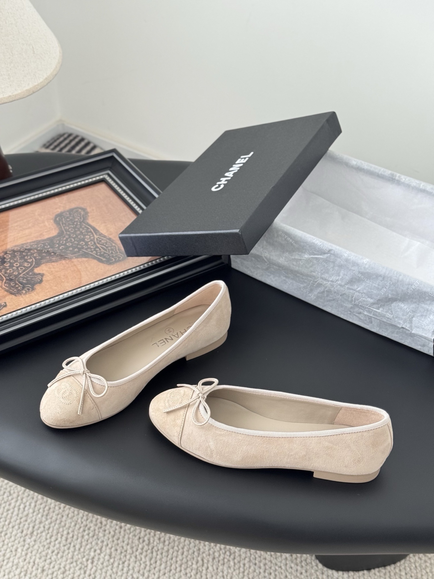 Pair of beige Chanel suede ballet flats with embossed CC logo and bow detail next to a black Chanel shoe box. - Hình 2