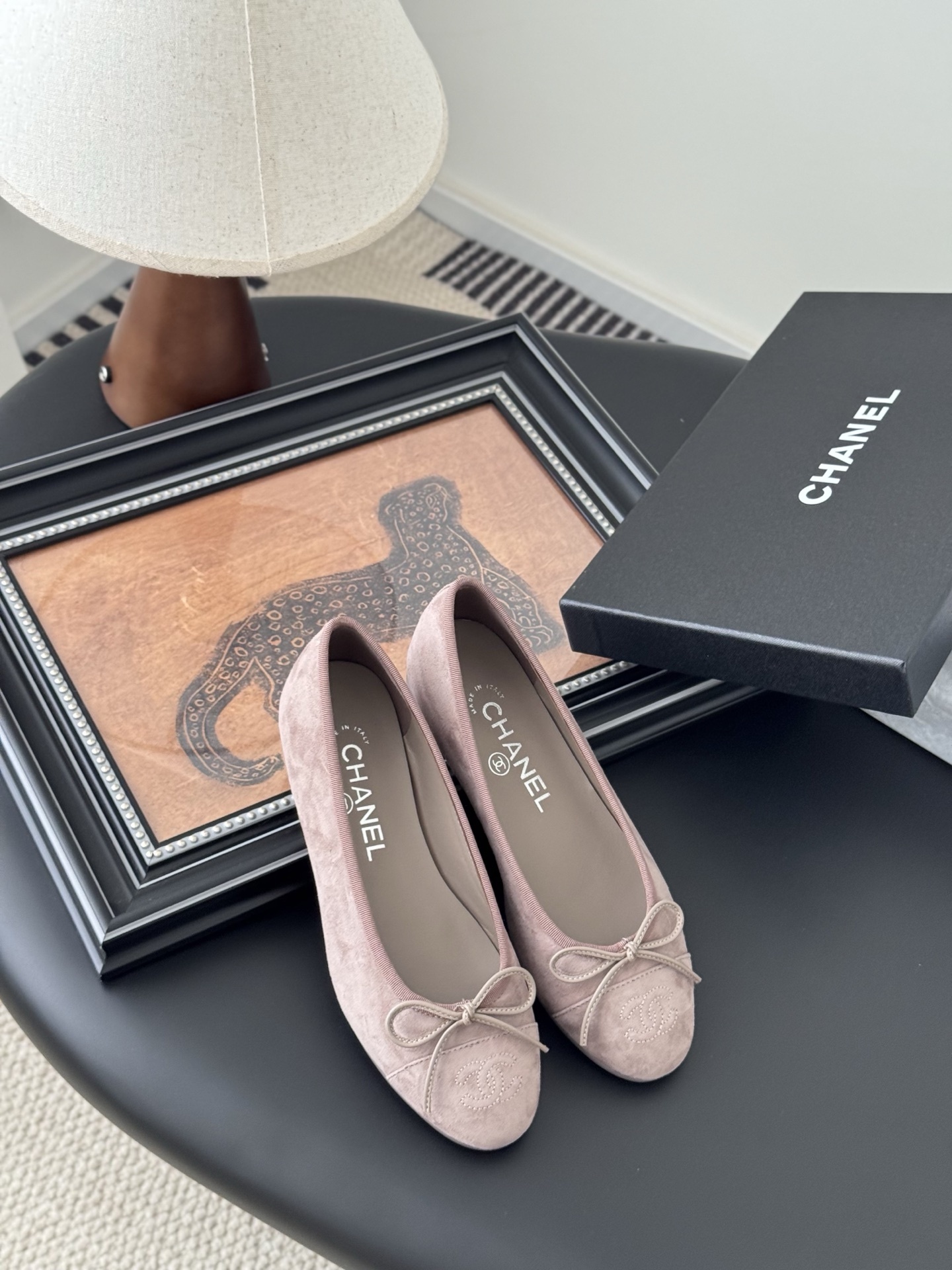 A pair of luxury pink Chanel suede ballet flats with bow and CC logo next to a black Chanel box on a black surface. - Hình 7