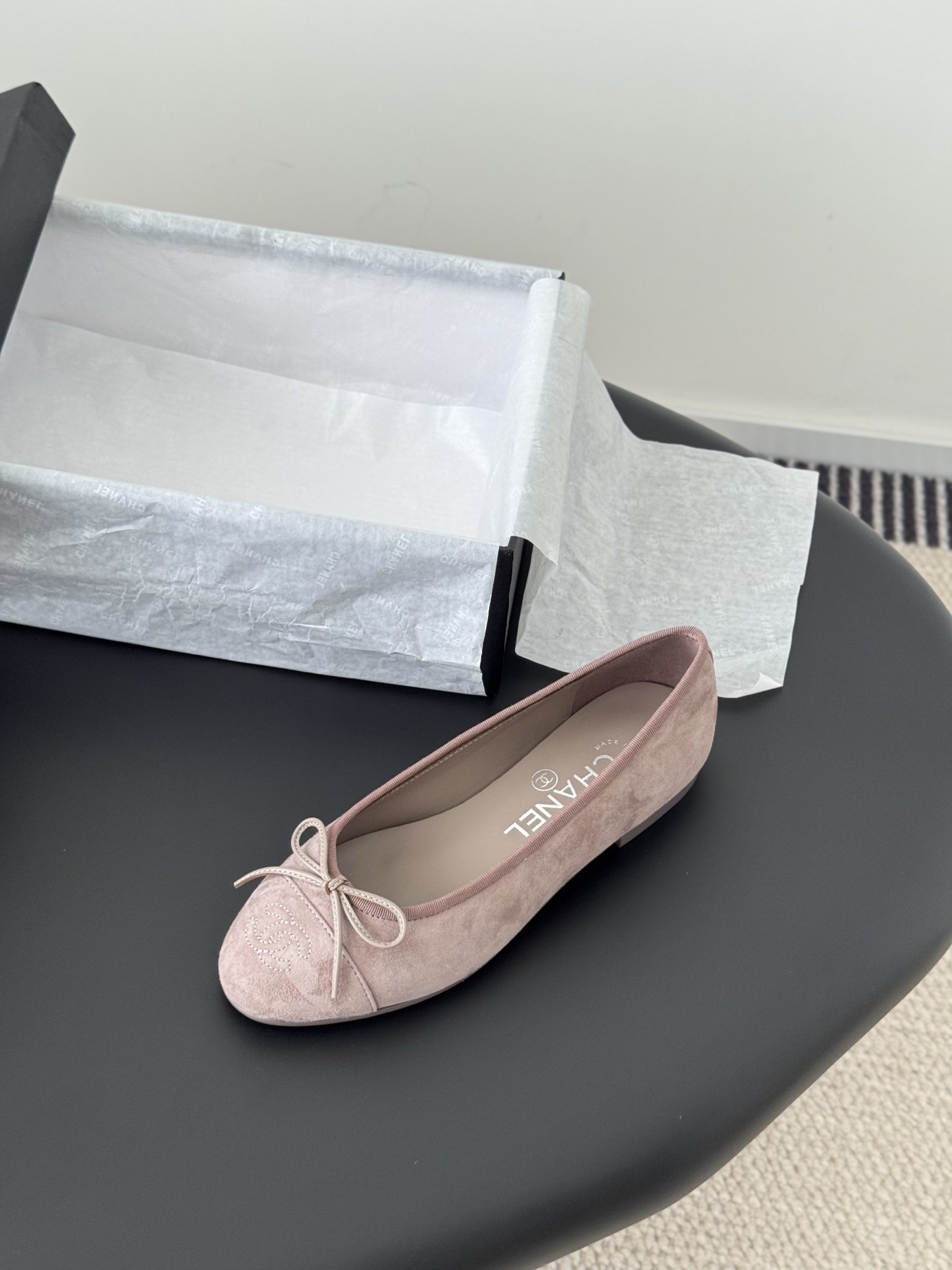 A pair of luxury pink Chanel suede ballet flats with bow and CC logo next to a black Chanel box on a black surface. - Hình 5