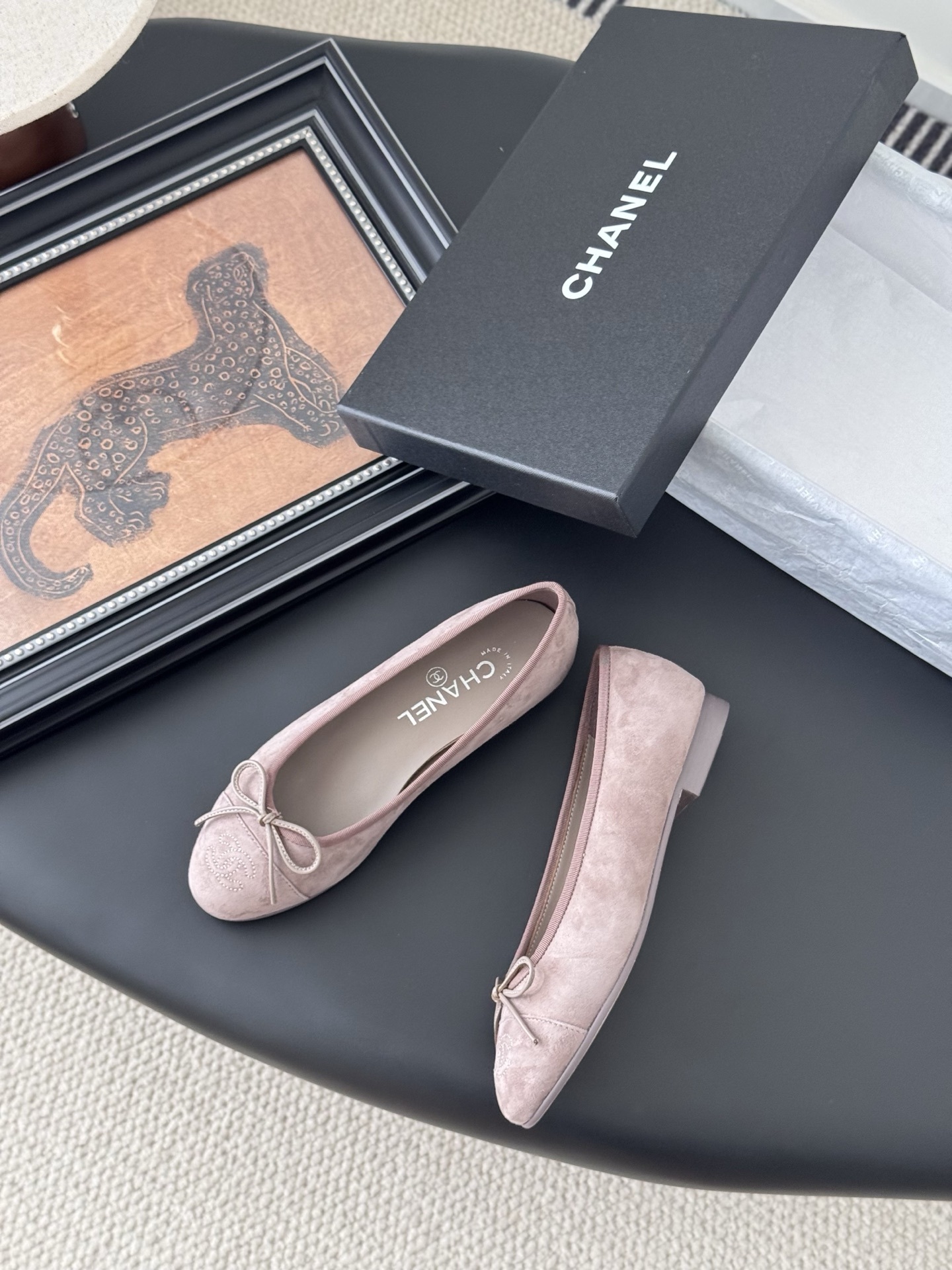 A pair of luxury pink Chanel suede ballet flats with bow and CC logo next to a black Chanel box on a black surface. - Hình 3