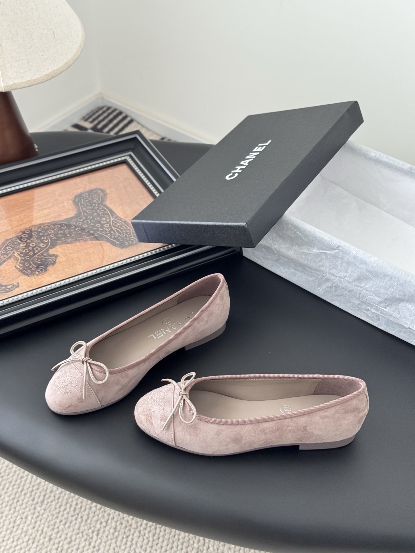 A pair of luxury pink Chanel suede ballet flats with bow and CC logo next to a black Chanel box on a black surface. - Hình 2