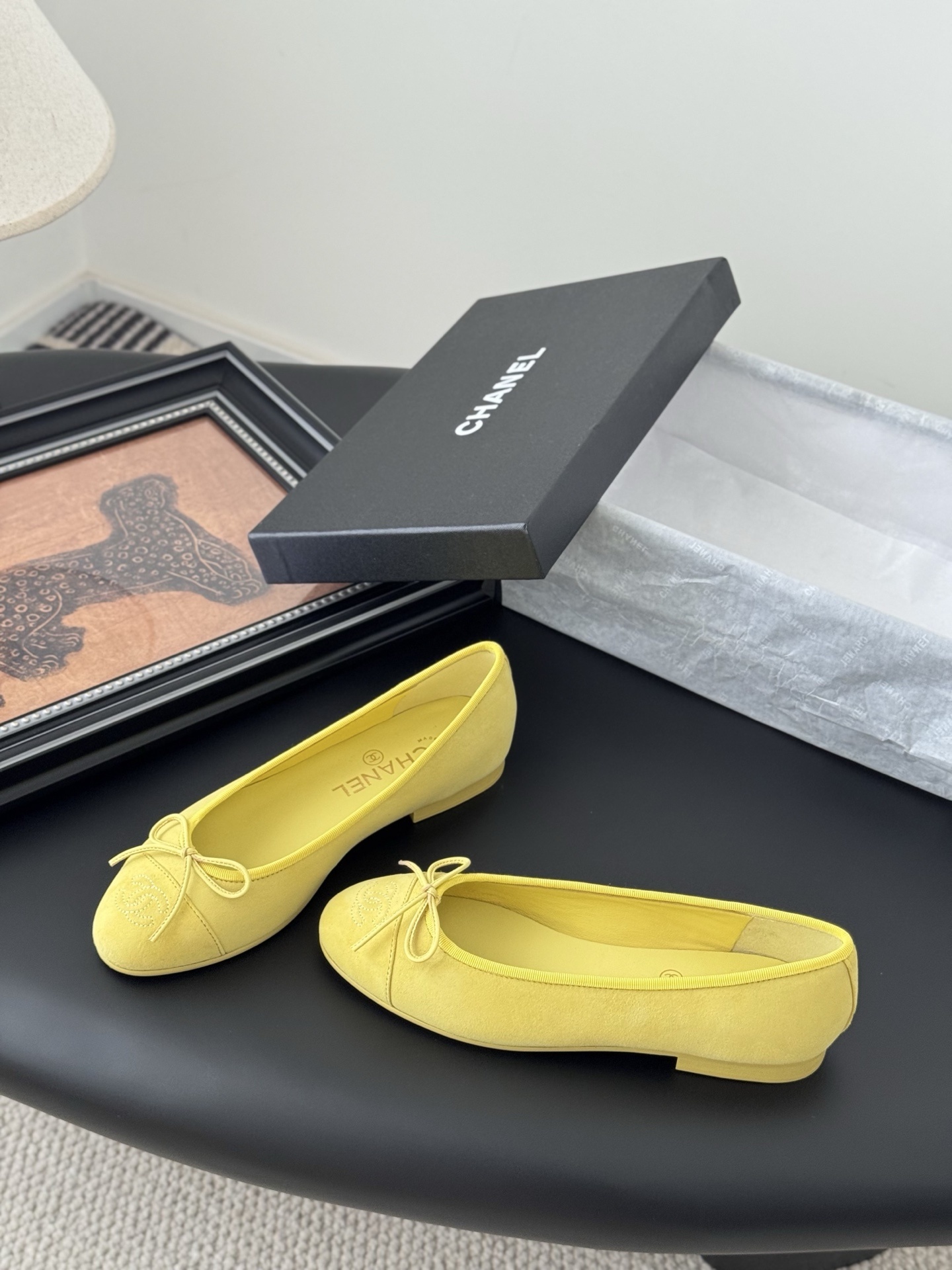 A pair of yellow Chanel suede ballerina flats with a bow and embroidered logo next to their original black box. - Hình 2