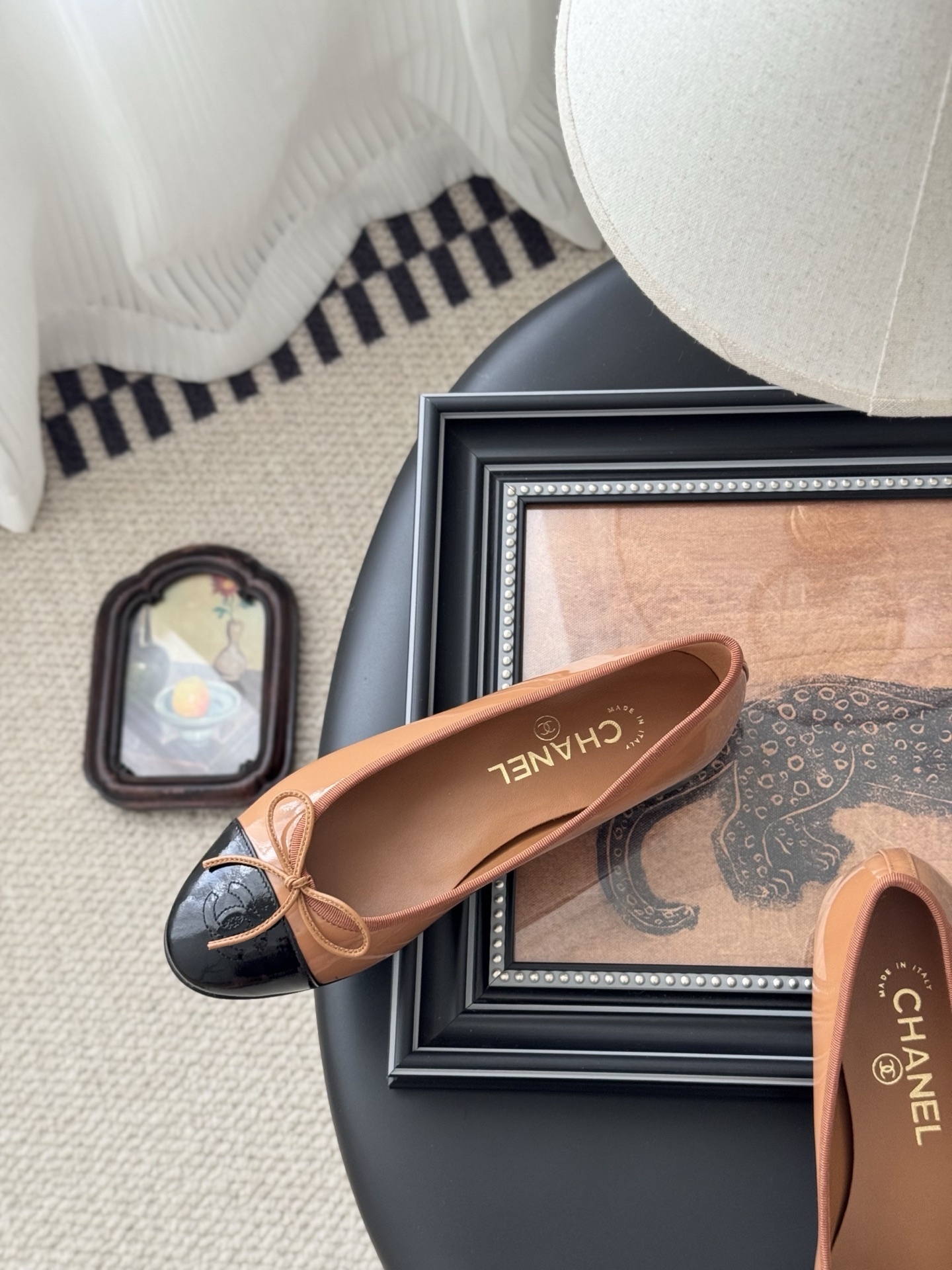 A pair of tan Chanel ballerina flats with black patent leather toe caps and bow details, placed next to a black Chanel shoe box. - Hình 8