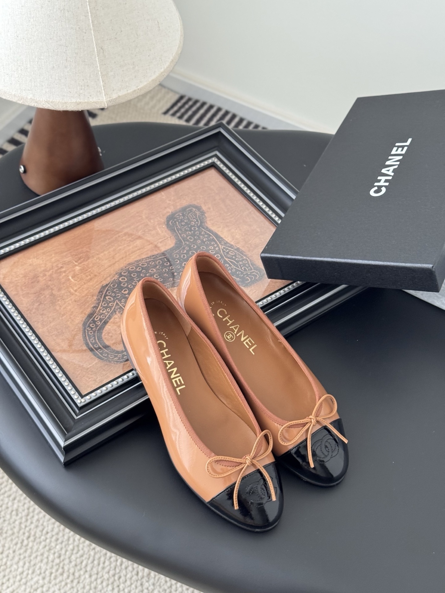 A pair of tan Chanel ballerina flats with black patent leather toe caps and bow details, placed next to a black Chanel shoe box. - Hình 7