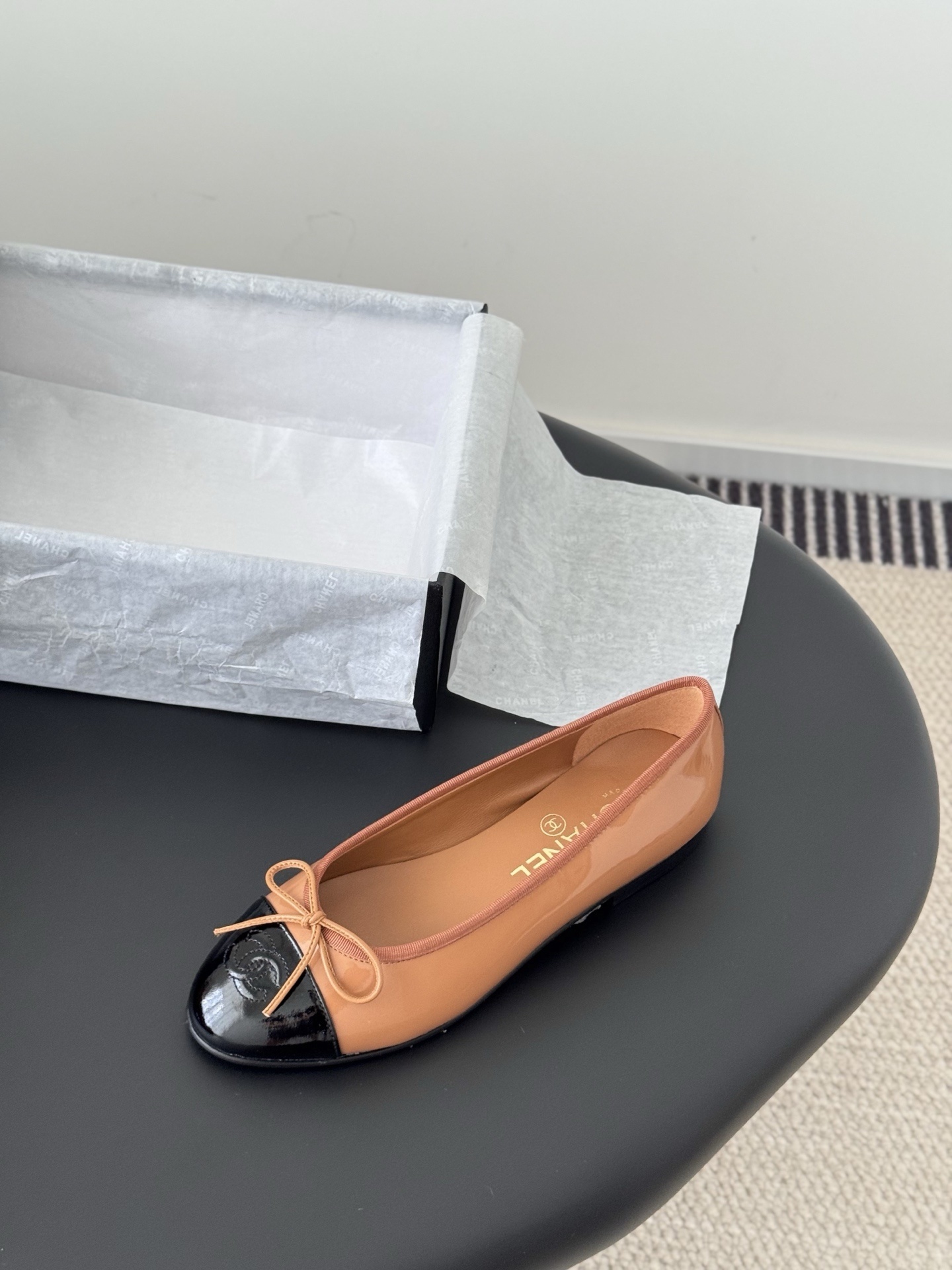 A pair of tan Chanel ballerina flats with black patent leather toe caps and bow details, placed next to a black Chanel shoe box. - Hình 5