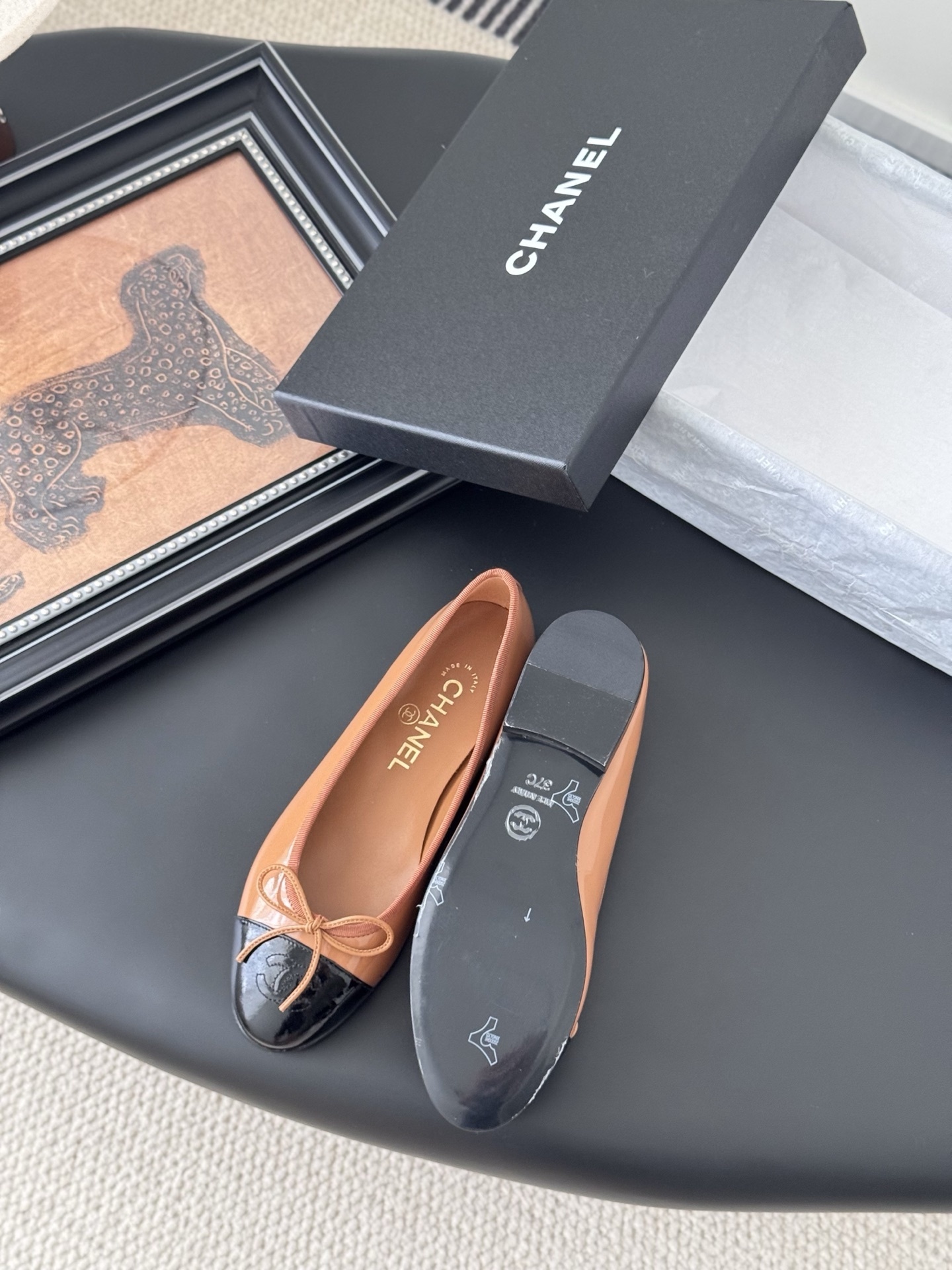 A pair of tan Chanel ballerina flats with black patent leather toe caps and bow details, placed next to a black Chanel shoe box. - Hình 4