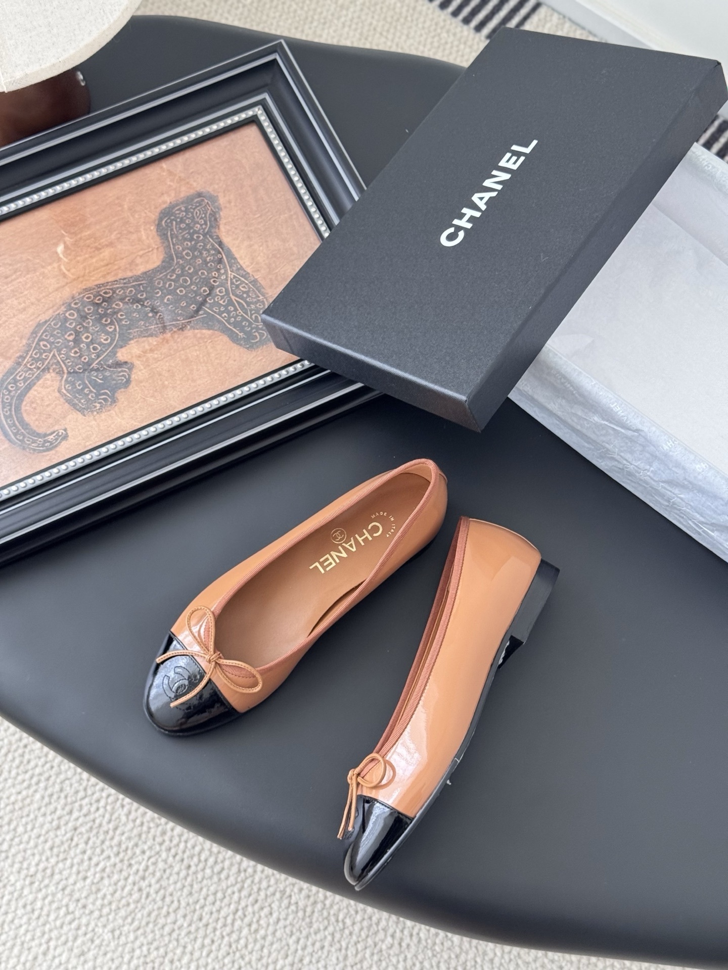 A pair of tan Chanel ballerina flats with black patent leather toe caps and bow details, placed next to a black Chanel shoe box. - Hình 3
