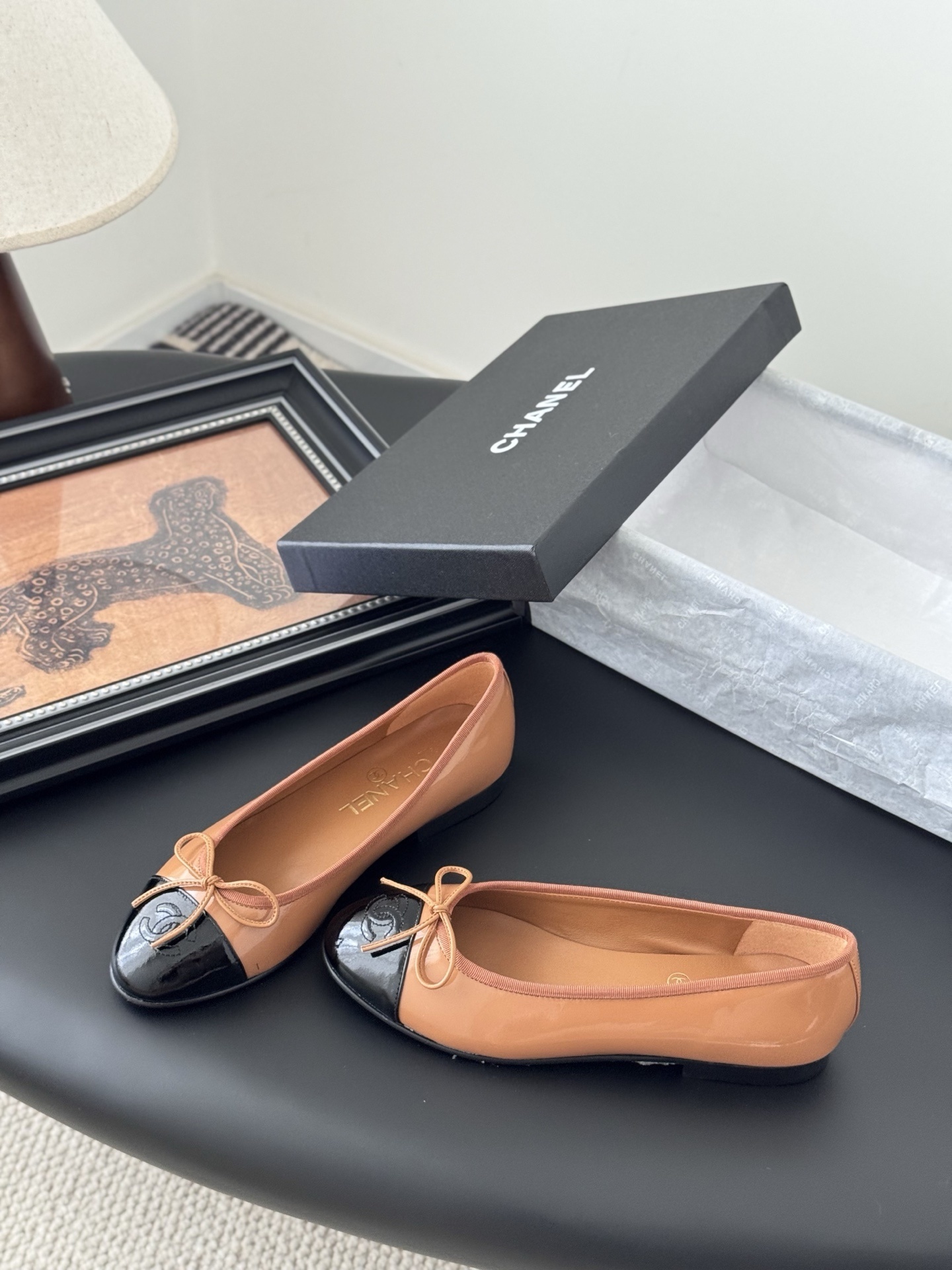 A pair of tan Chanel ballerina flats with black patent leather toe caps and bow details, placed next to a black Chanel shoe box. - Hình 2