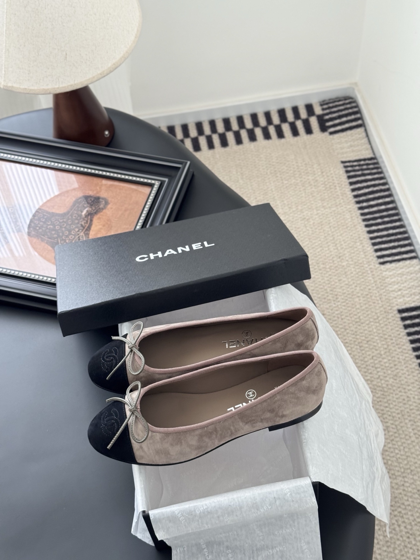 Luxury Chanel beige suede ballerina flats with black CC logo toe cap placed next to a black Chanel shoe box on a dark table. - Hình 9