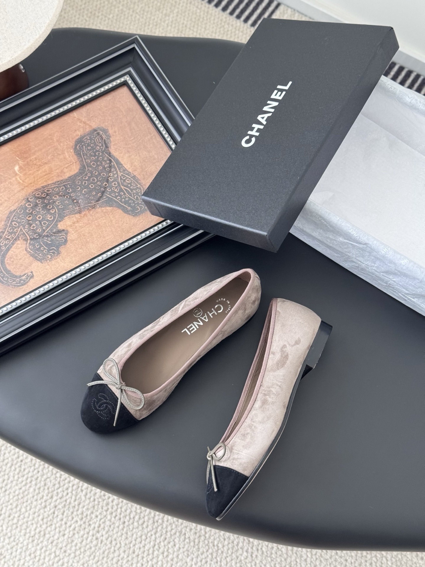 Luxury Chanel beige suede ballerina flats with black CC logo toe cap placed next to a black Chanel shoe box on a dark table. - Hình 3