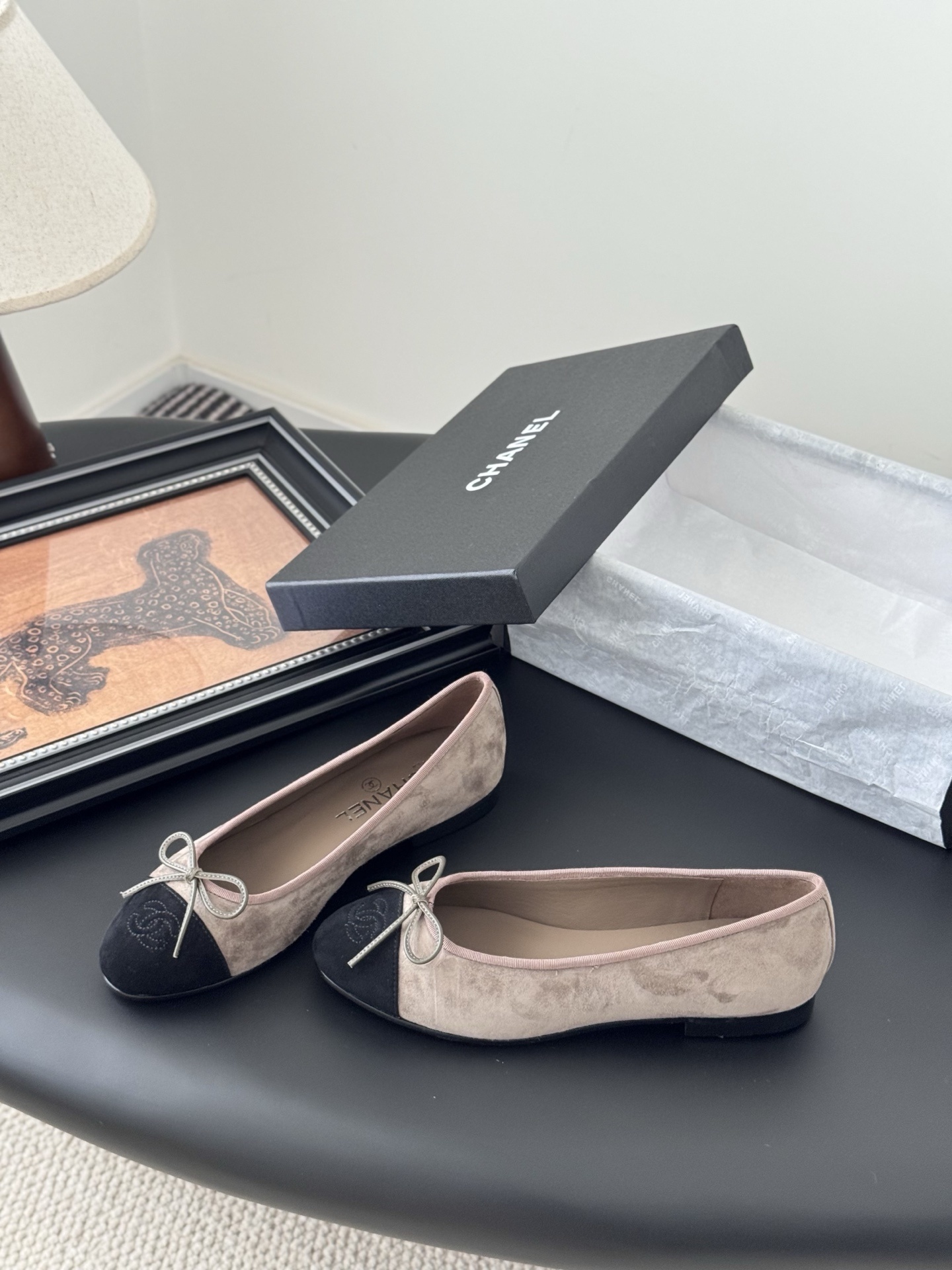Luxury Chanel beige suede ballerina flats with black CC logo toe cap placed next to a black Chanel shoe box on a dark table. - Hình 2