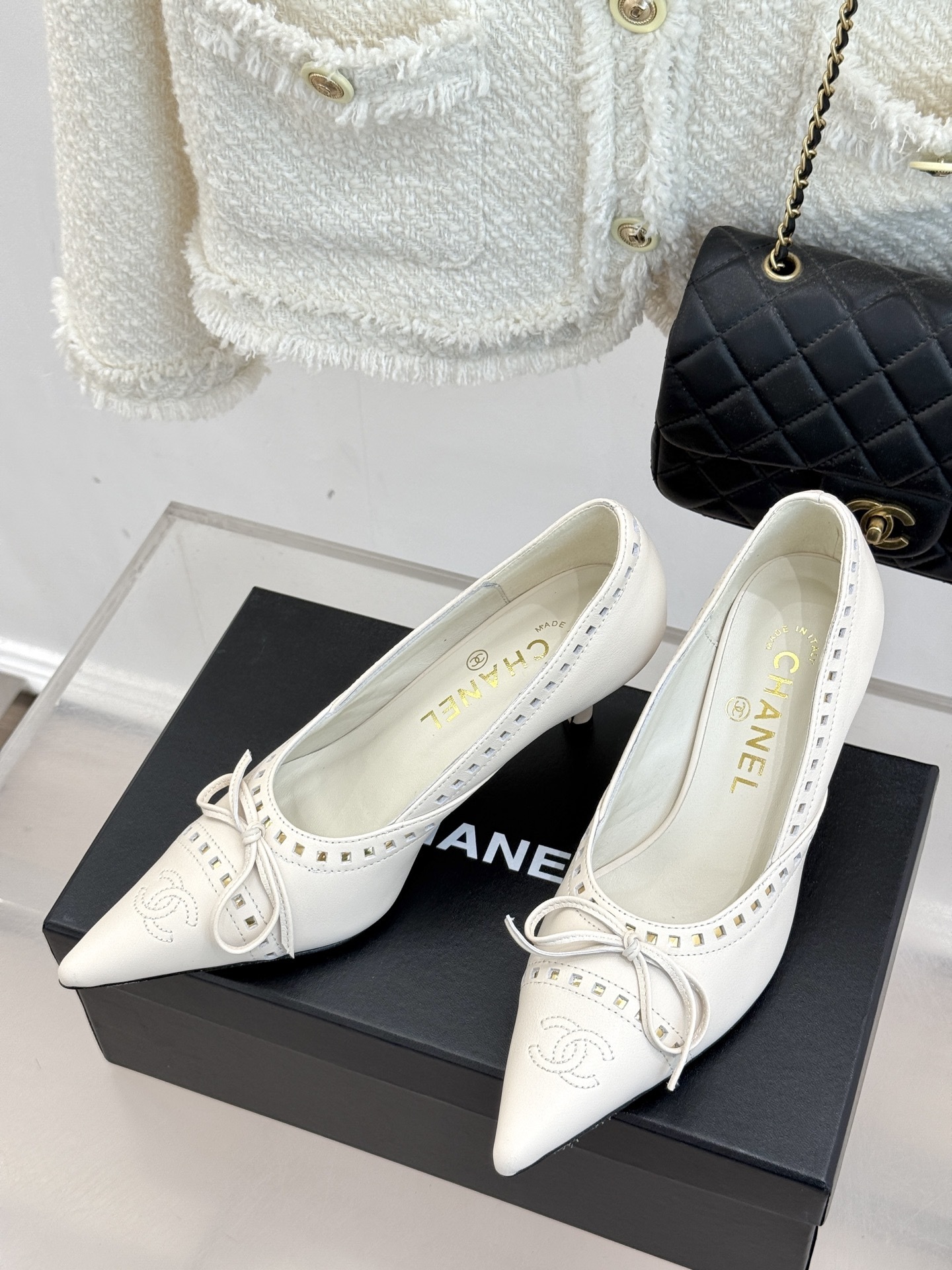 Luxury Chanel white leather pointed-toe kitten heels with bow and embossed CC logo on a black shoe box. - Hình 2