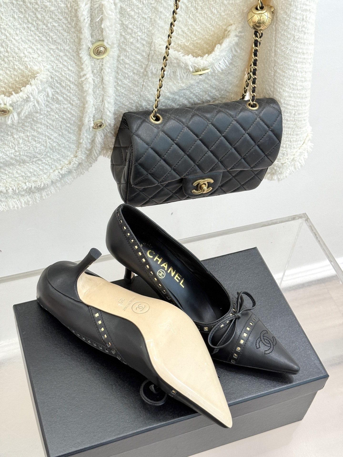 A pair of luxury black Chanel leather pointed-toe pumps with gold stud details and a bow, resting on a black shoe box. - Hình 9