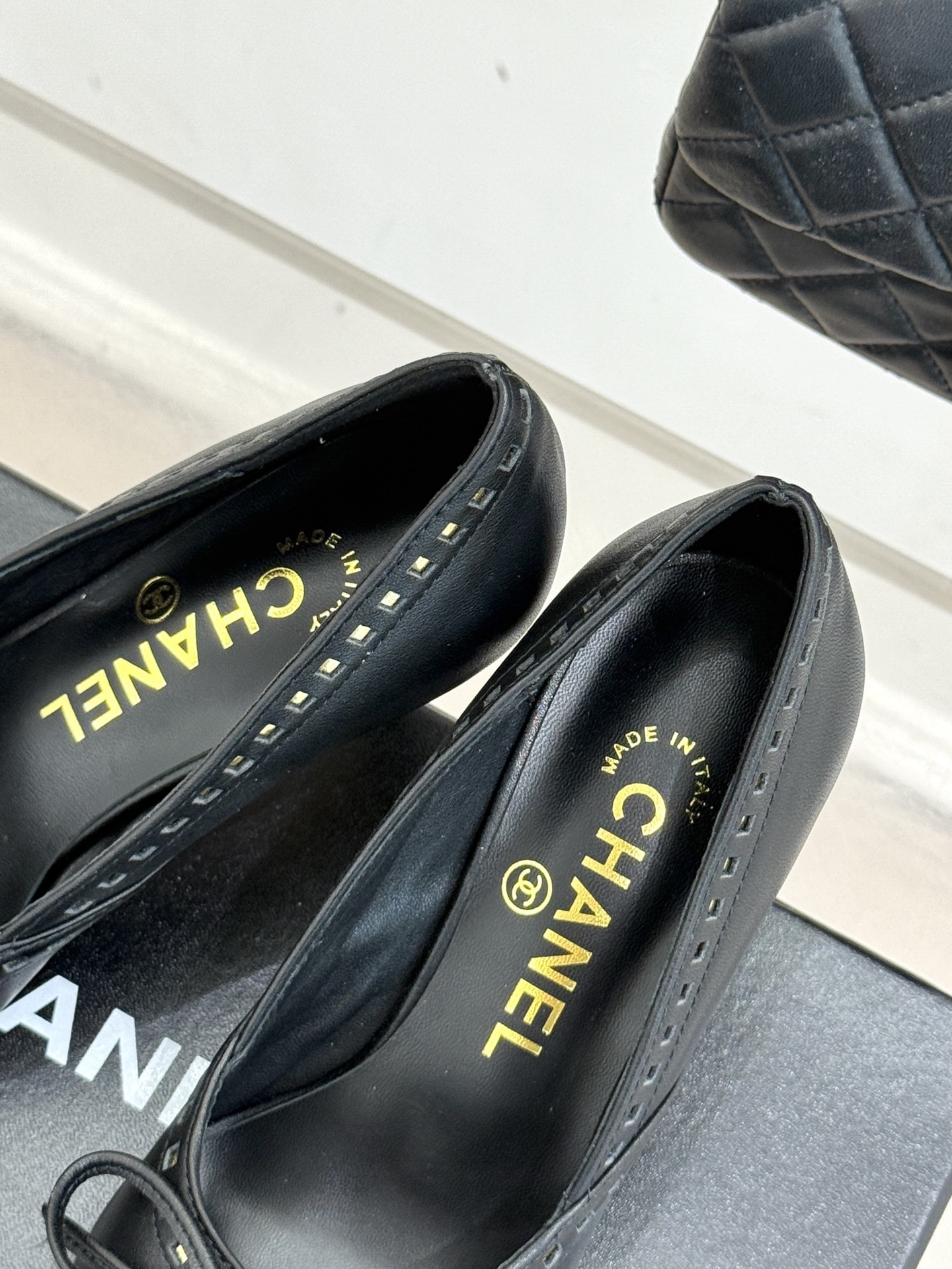 A pair of luxury black Chanel leather pointed-toe pumps with gold stud details and a bow, resting on a black shoe box. - Hình 8