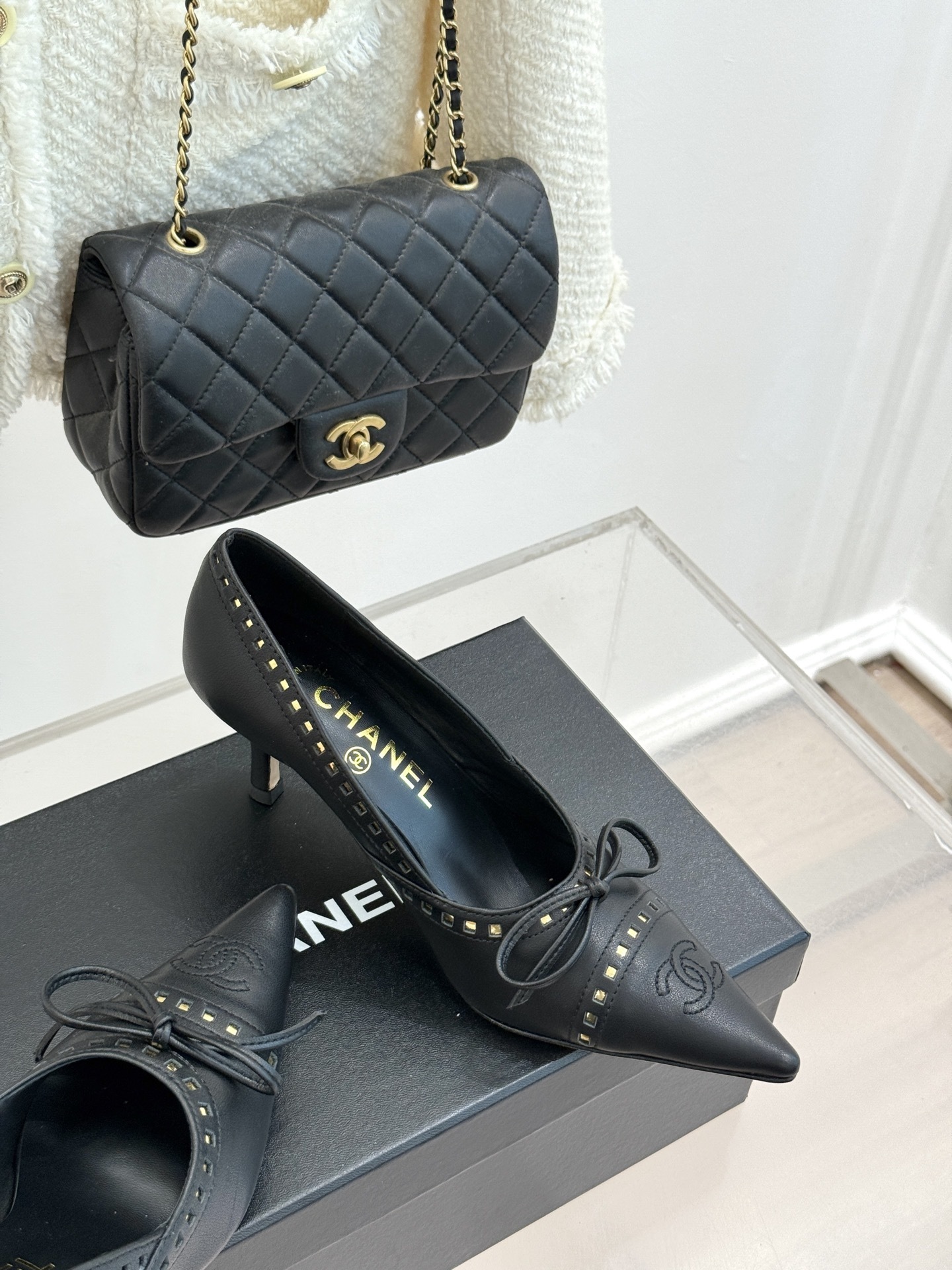 A pair of luxury black Chanel leather pointed-toe pumps with gold stud details and a bow, resting on a black shoe box. - Hình 5