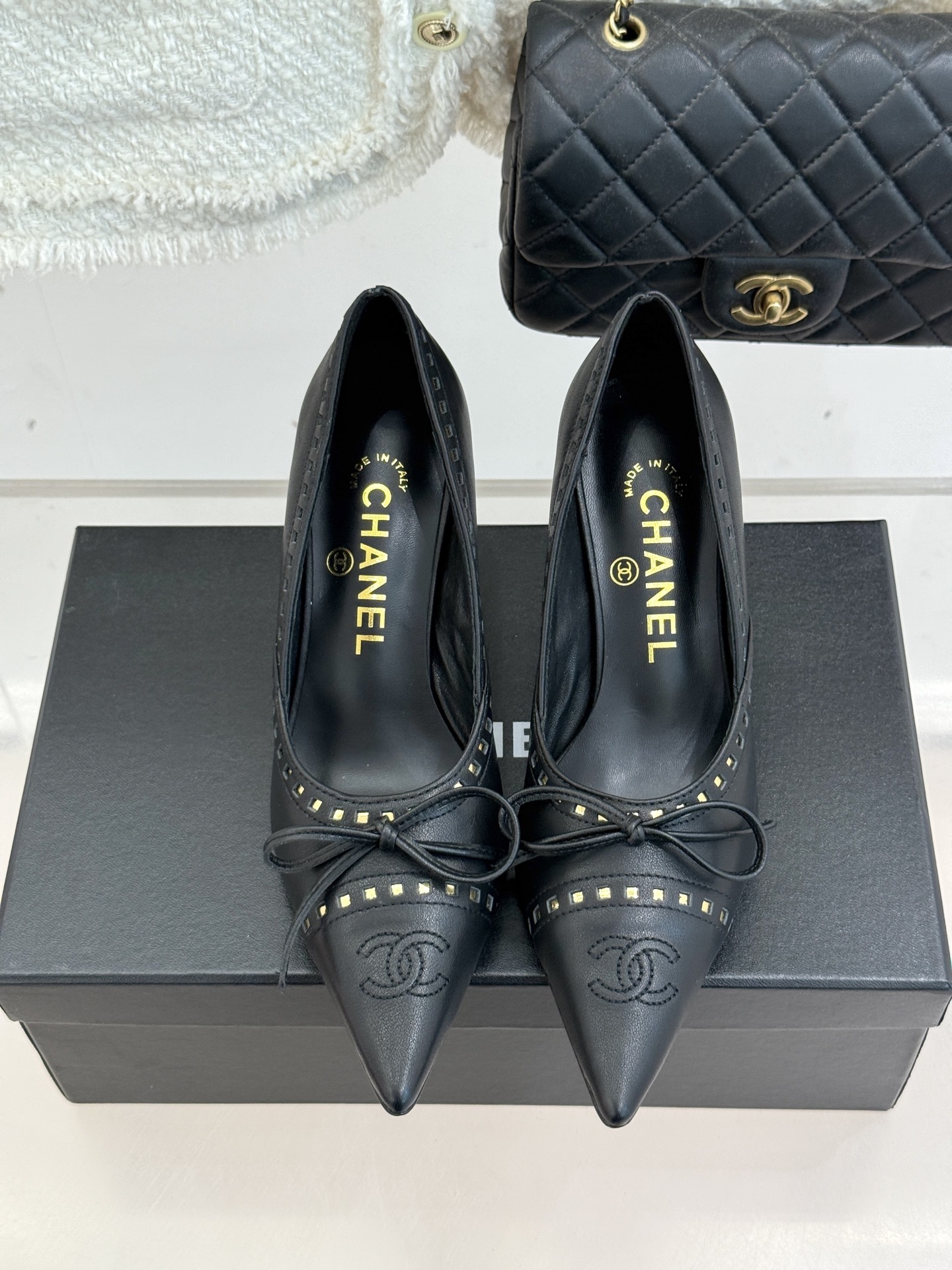 A pair of luxury black Chanel leather pointed-toe pumps with gold stud details and a bow, resting on a black shoe box. - Hình 4