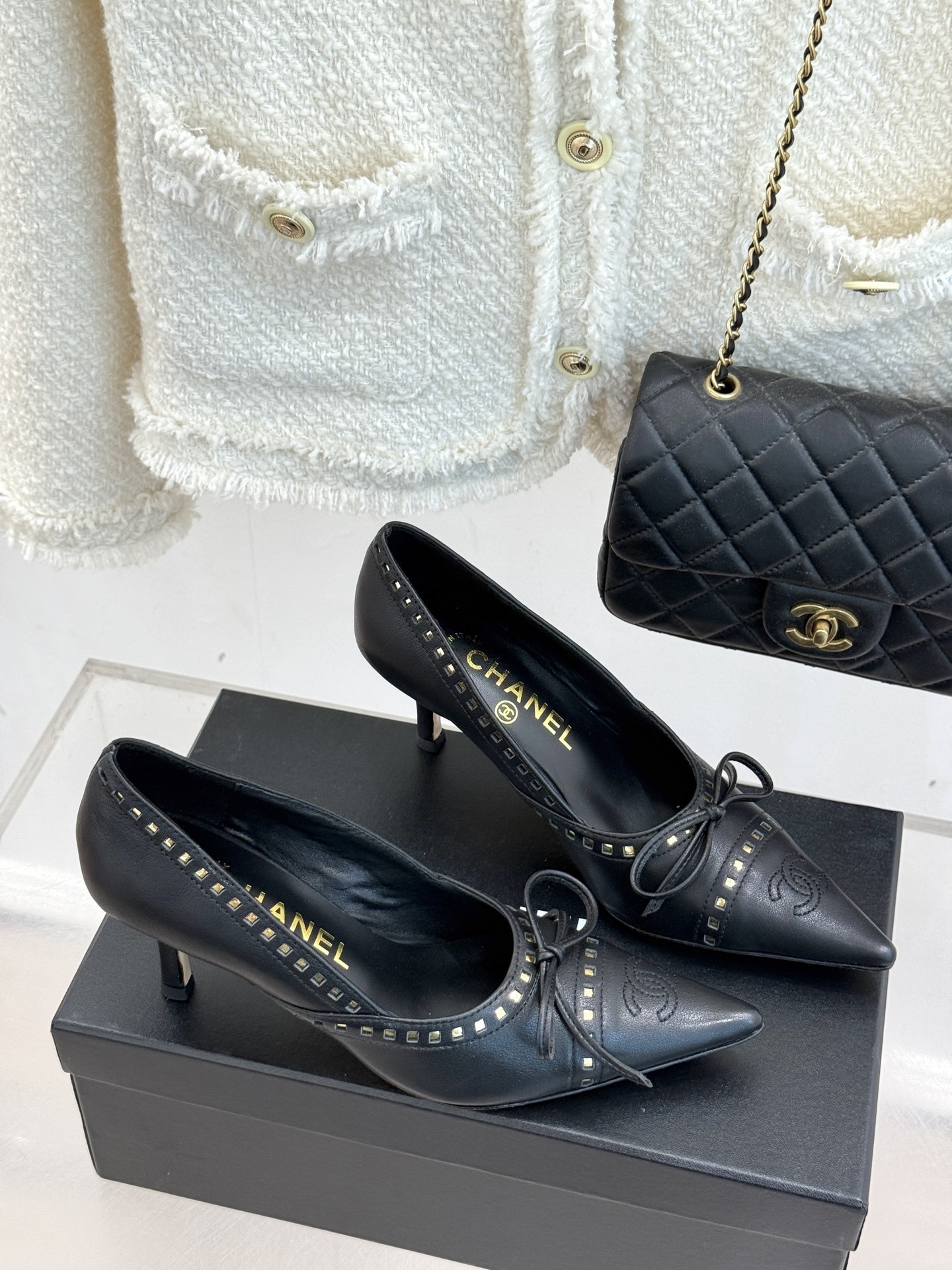 A pair of luxury black Chanel leather pointed-toe pumps with gold stud details and a bow, resting on a black shoe box. - Hình 3