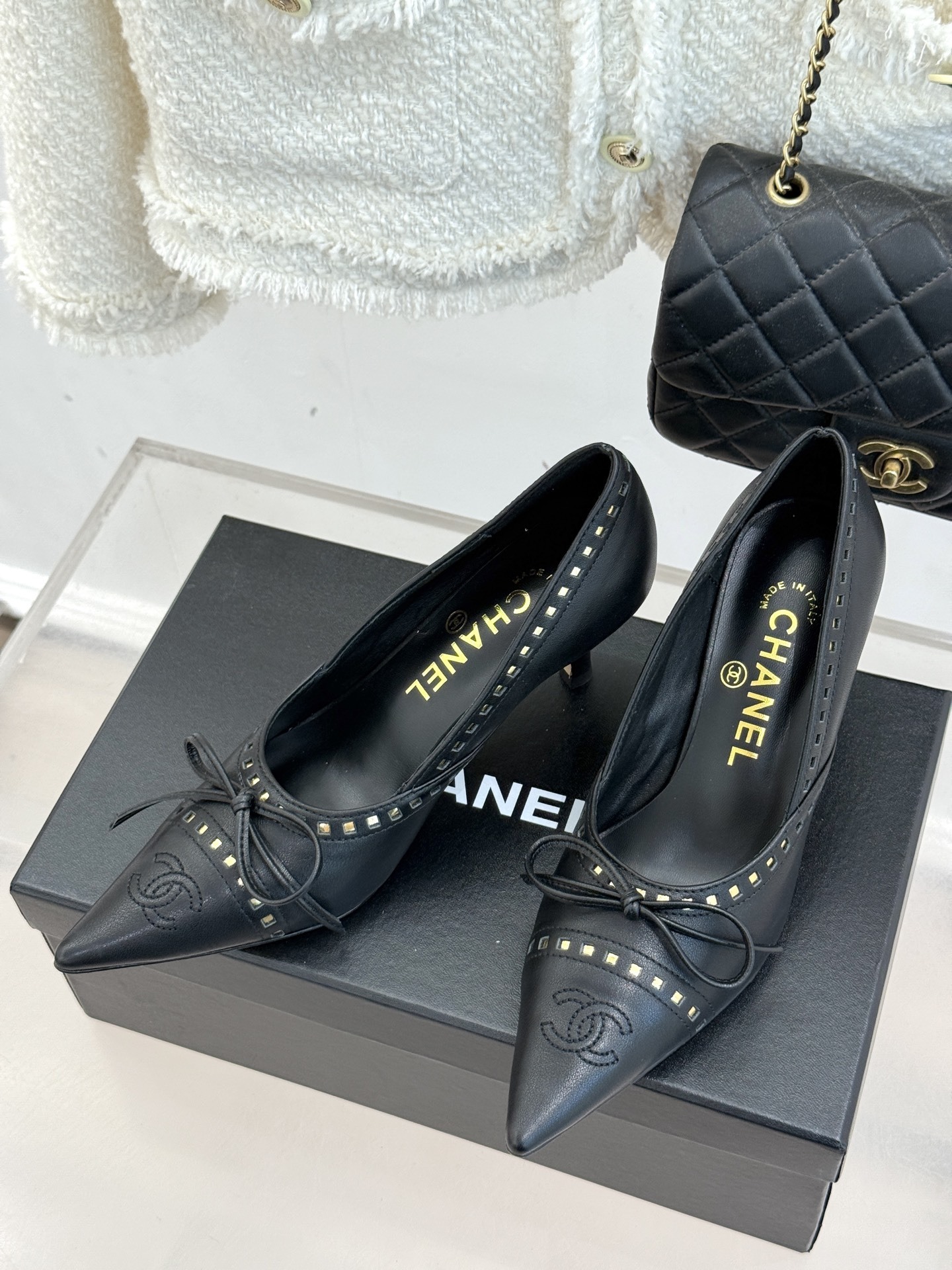 A pair of luxury black Chanel leather pointed-toe pumps with gold stud details and a bow, resting on a black shoe box. - Hình 2
