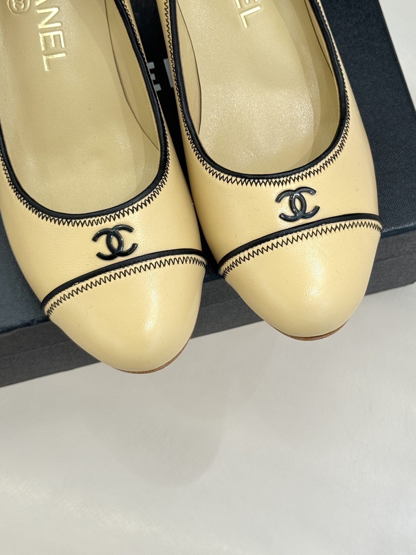 A pair of beige Chanel kitten heel pumps with black trim and CC logo on a black shoebox - Hình 7