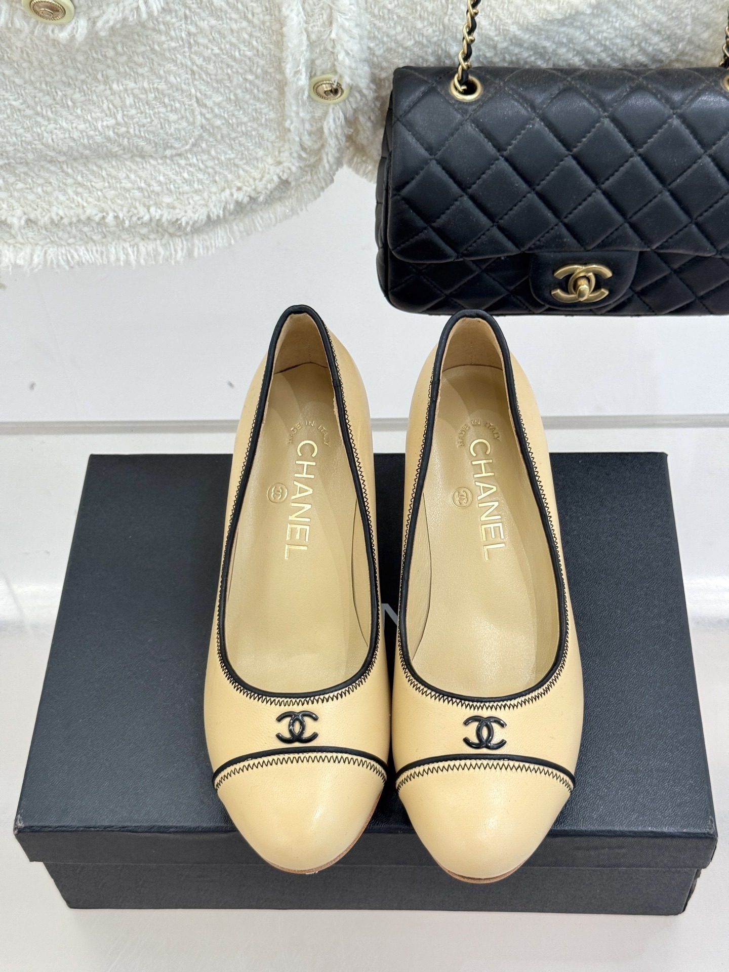 A pair of beige Chanel kitten heel pumps with black trim and CC logo on a black shoebox - Hình 4