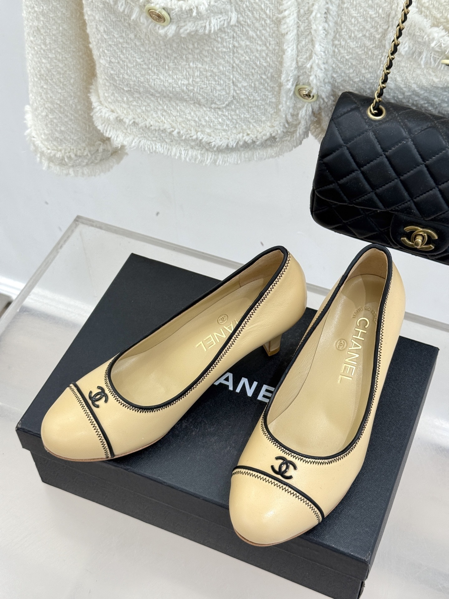 A pair of beige Chanel kitten heel pumps with black trim and CC logo on a black shoebox - Hình 2