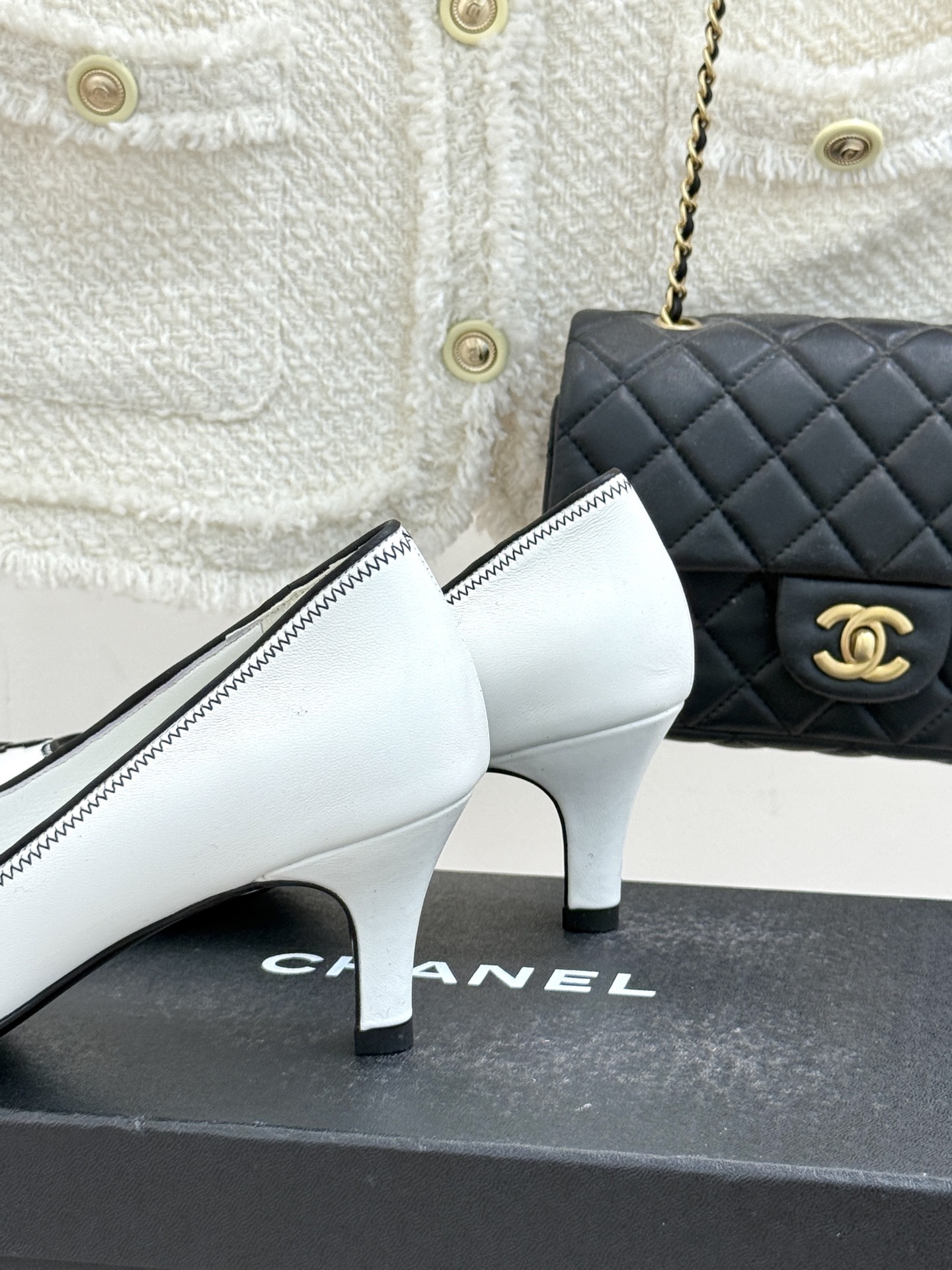 Elegant white Chanel kitten heels with black trim and CC logo on a black shoe box next to a quilted bag. - Hình 6