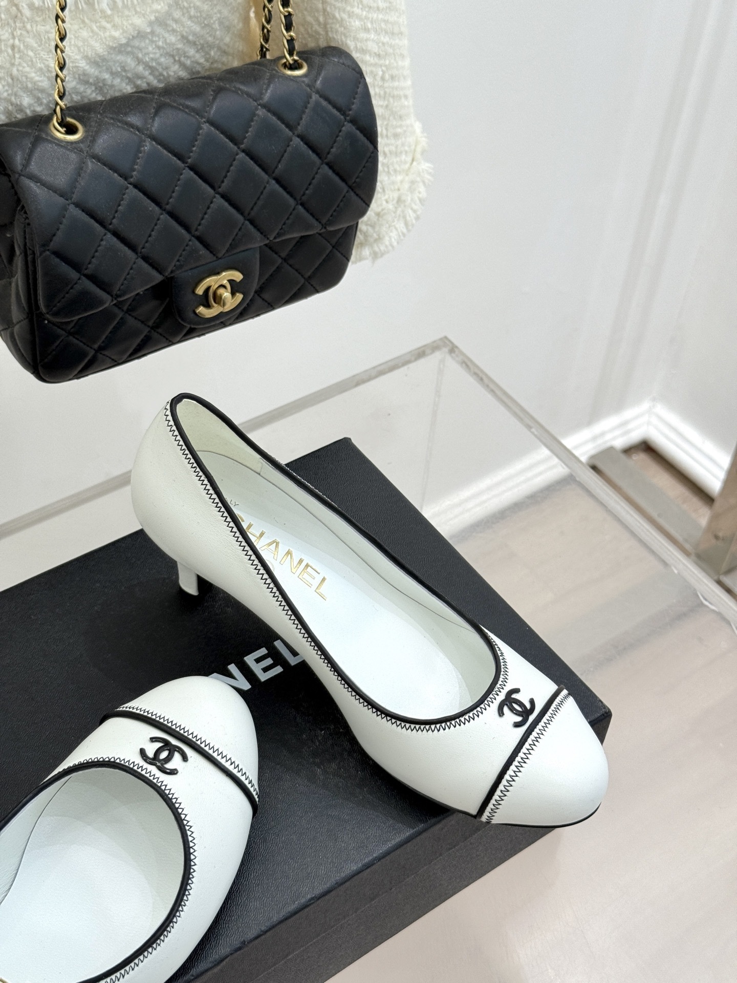 Elegant white Chanel kitten heels with black trim and CC logo on a black shoe box next to a quilted bag. - Hình 5