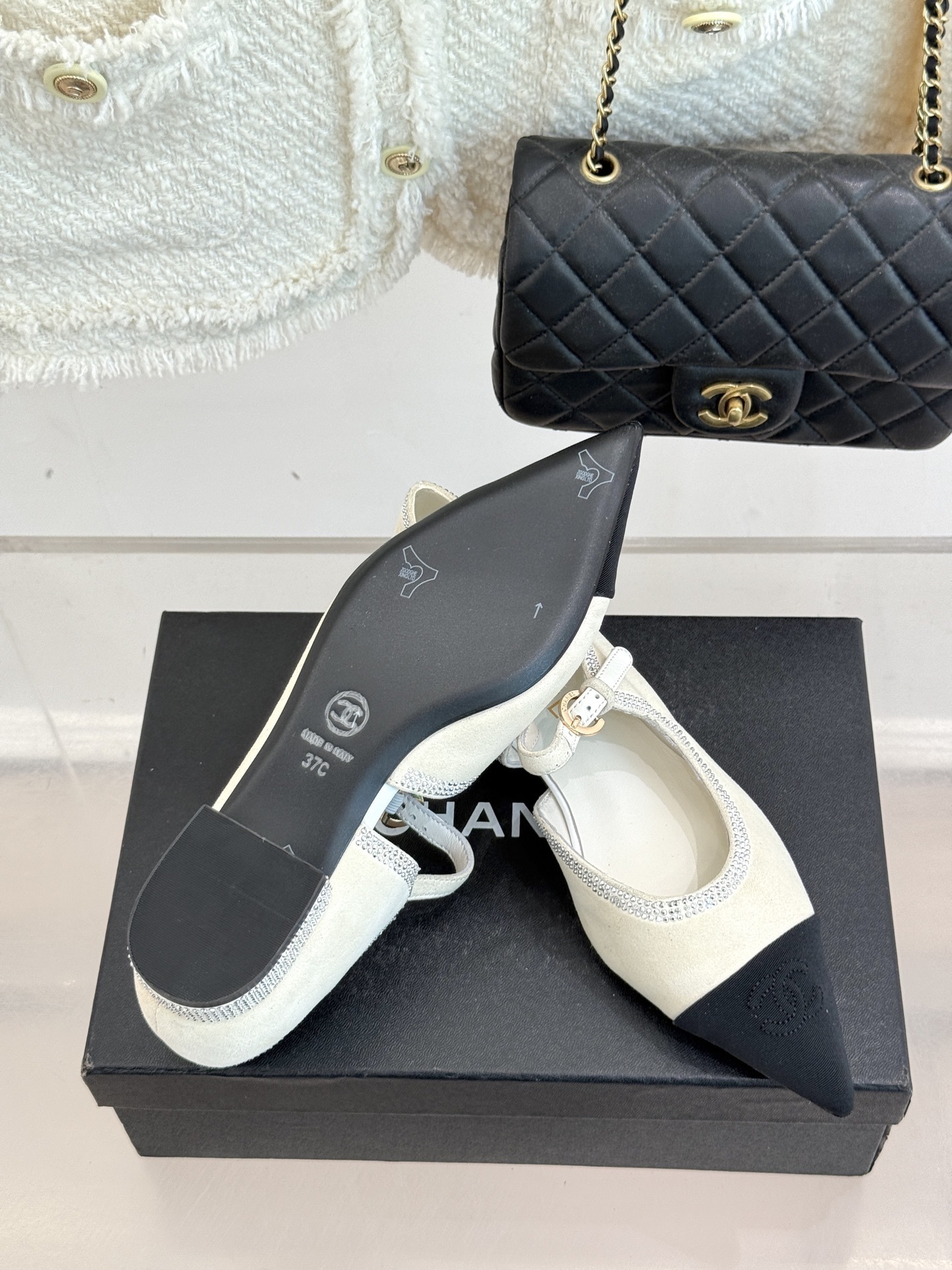 Luxury Chanel white suede Mary Jane flats with crystal trim and black cap toe displayed on a black designer box. - Hình 9