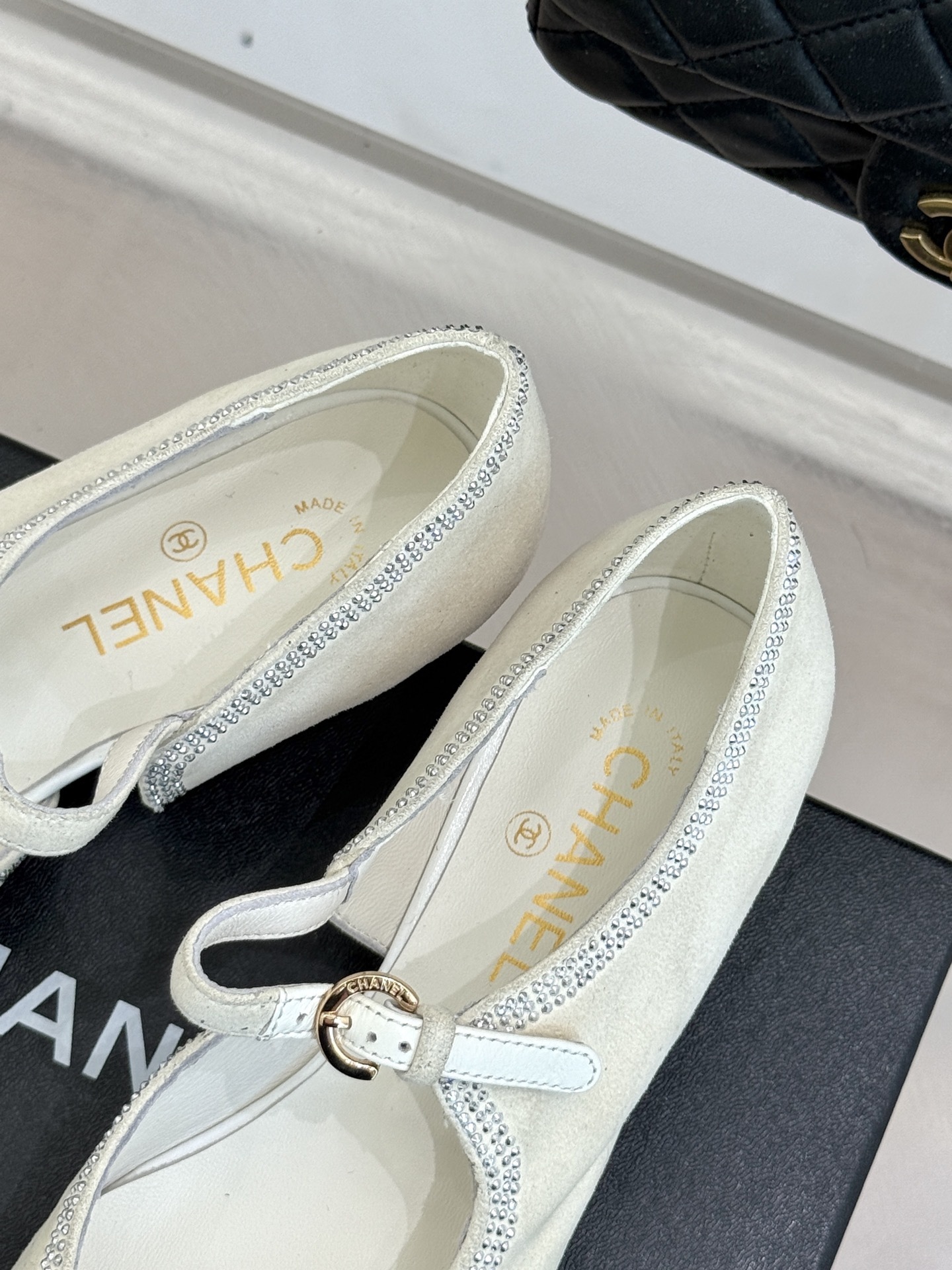 Luxury Chanel white suede Mary Jane flats with crystal trim and black cap toe displayed on a black designer box. - Hình 8
