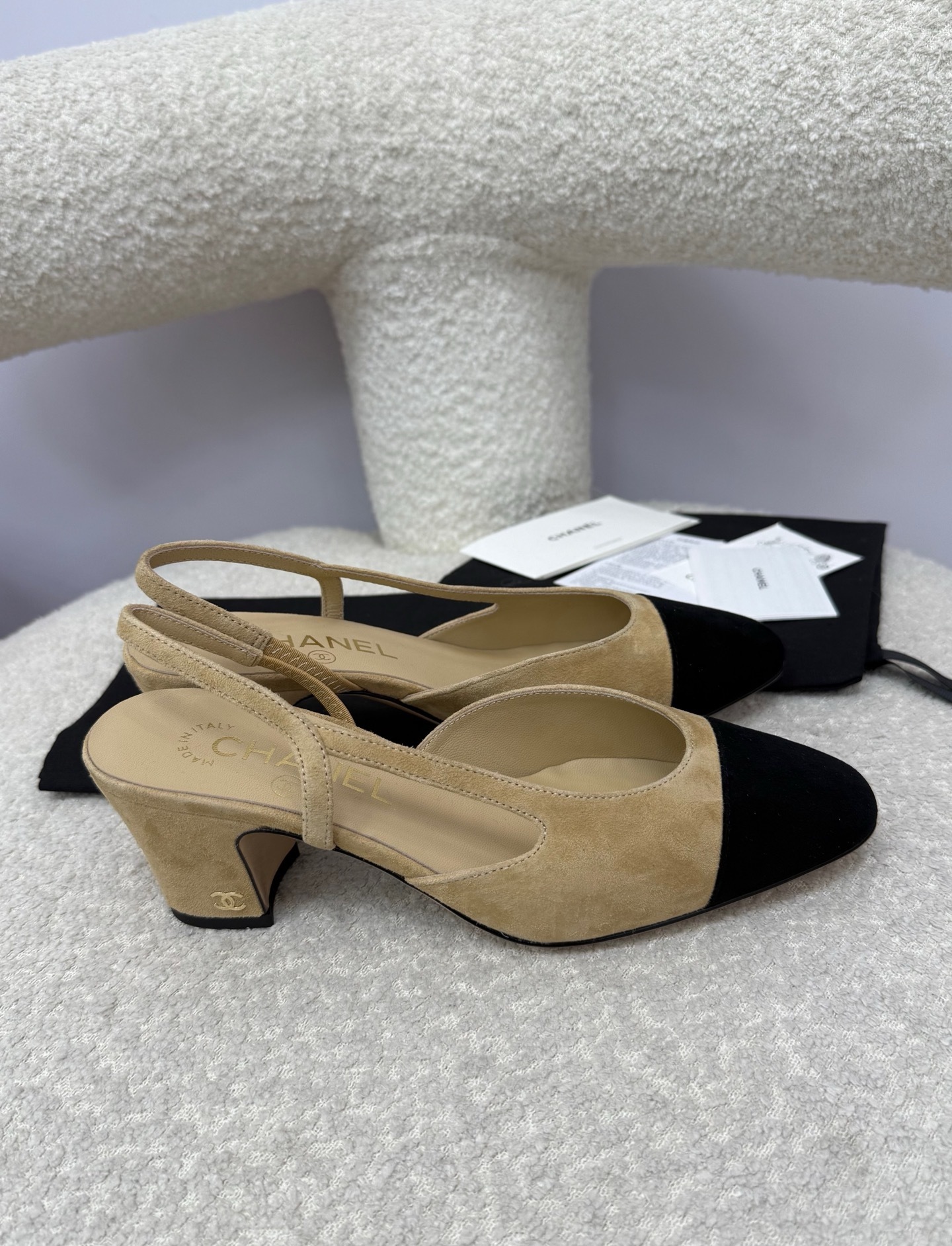 A pair of luxury Chanel beige suede slingback pumps with black cap toes and block heels on a white textured surface. - Hình 8