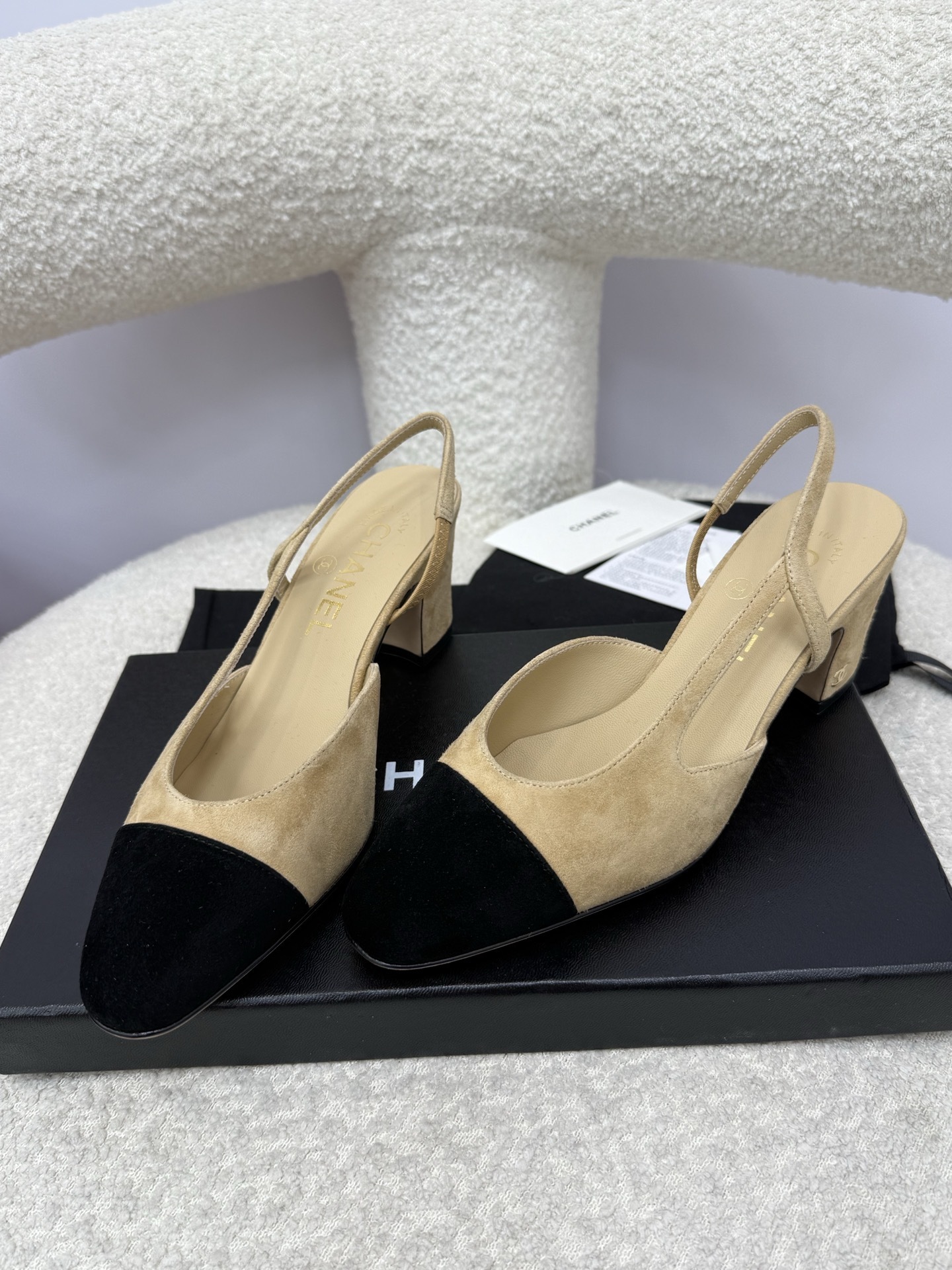 A pair of luxury Chanel beige suede slingback pumps with black cap toes and block heels on a white textured surface. - Hình 5