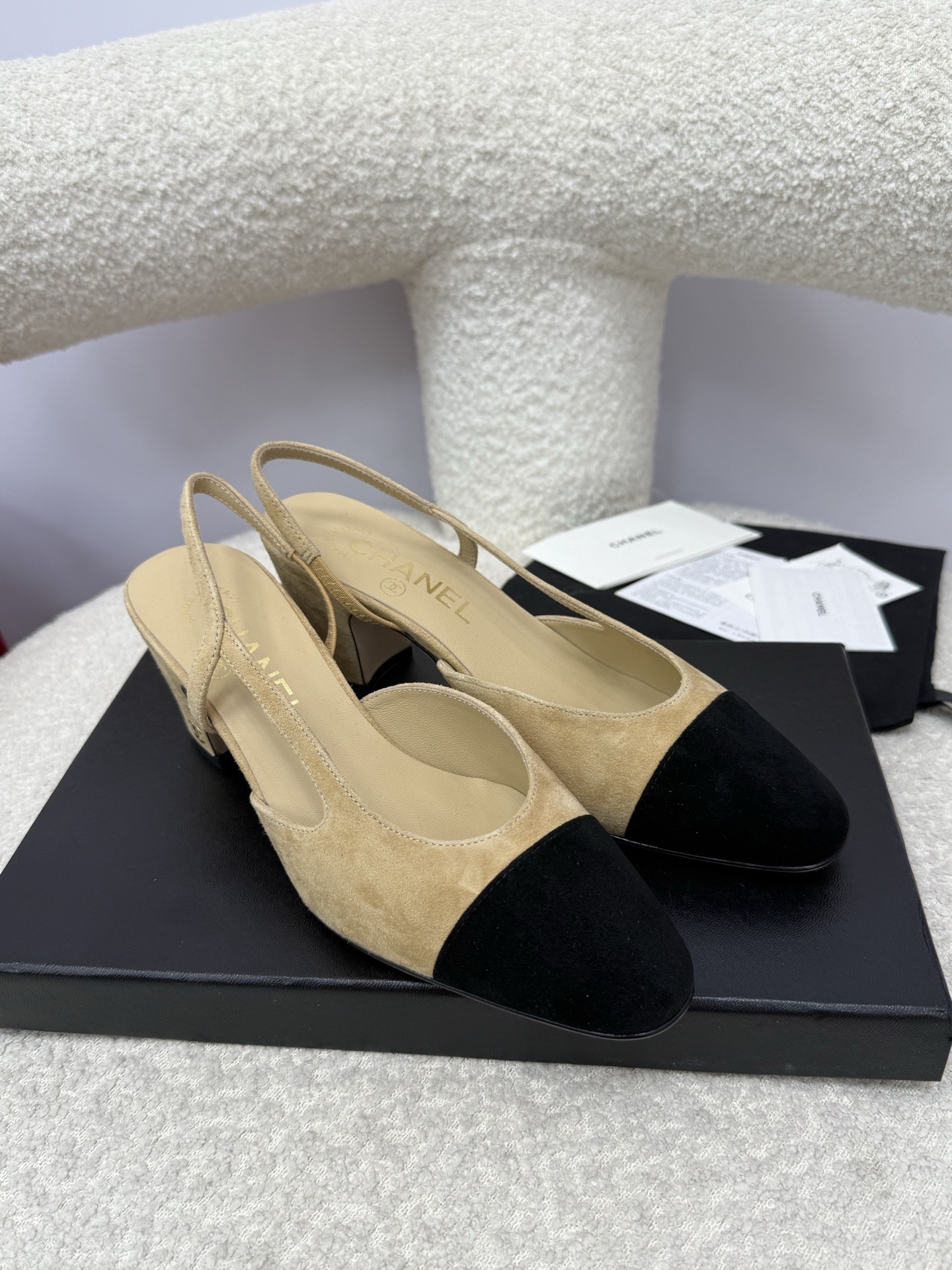 A pair of luxury Chanel beige suede slingback pumps with black cap toes and block heels on a white textured surface. - Hình 4