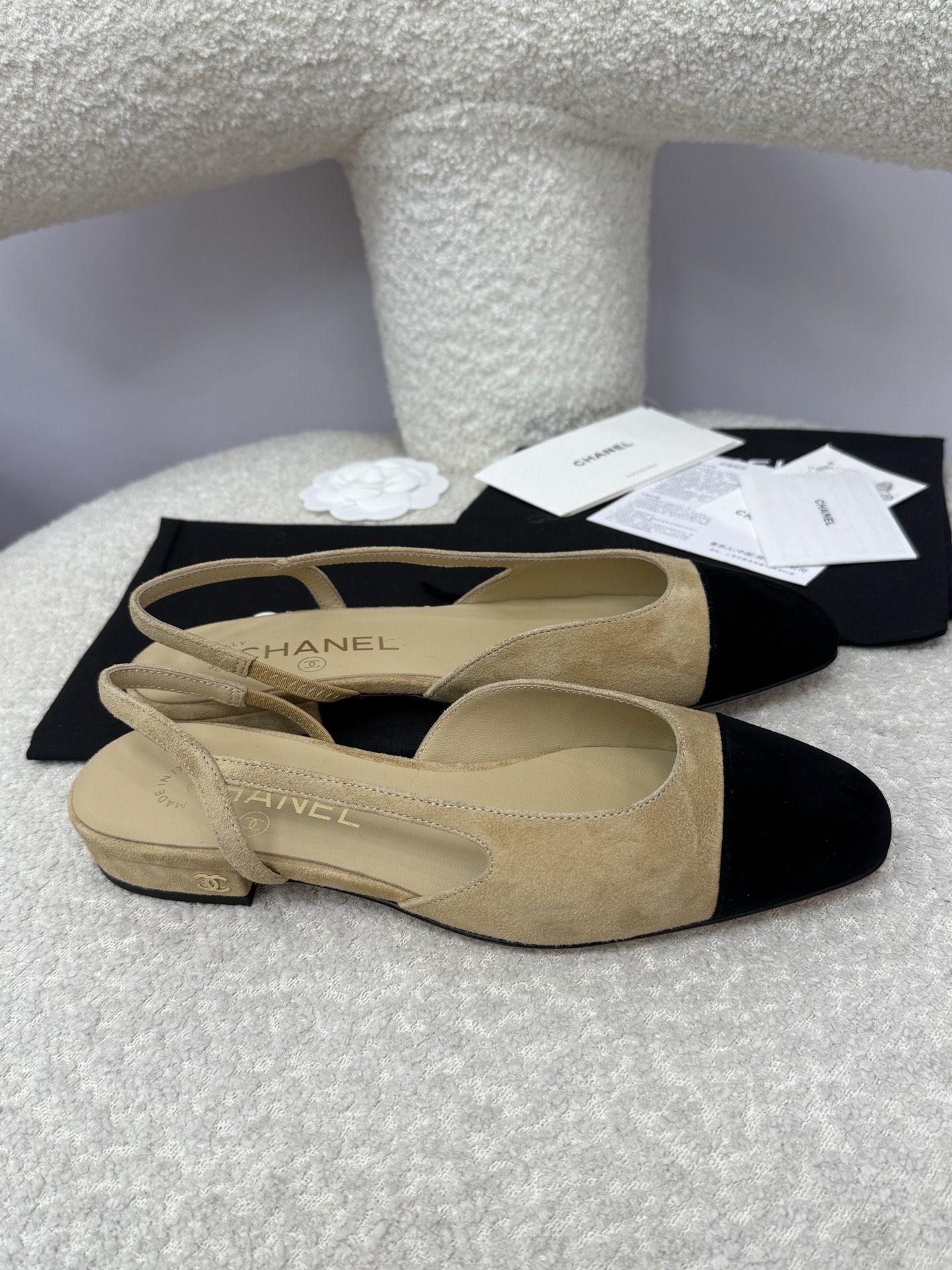 A pair of Chanel two-tone beige and black suede slingback flats displayed on a white textured surface with branded dust bags and authentication cards. - Hình 8