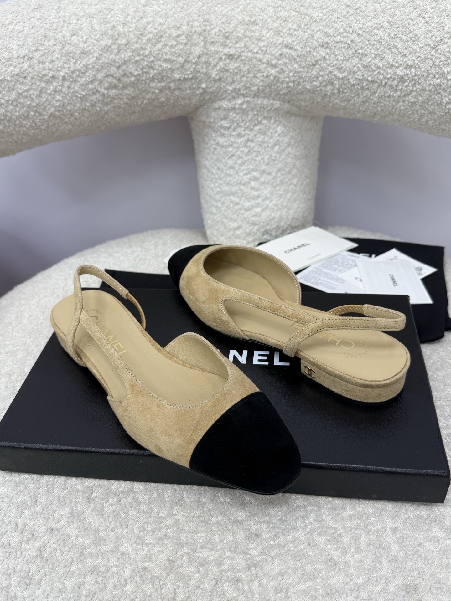A pair of Chanel two-tone beige and black suede slingback flats displayed on a white textured surface with branded dust bags and authentication cards. - Hình 7