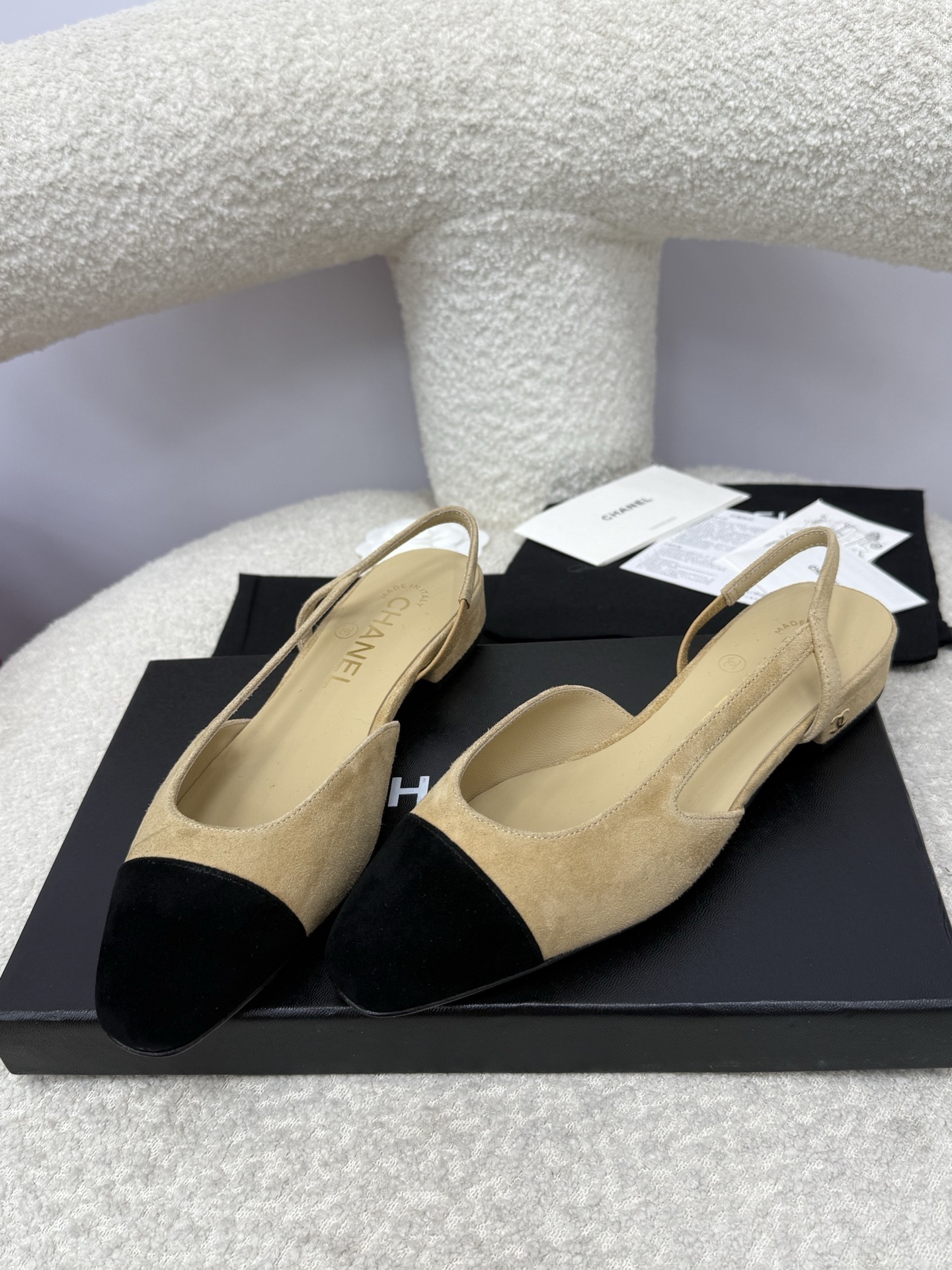 A pair of Chanel two-tone beige and black suede slingback flats displayed on a white textured surface with branded dust bags and authentication cards. - Hình 6