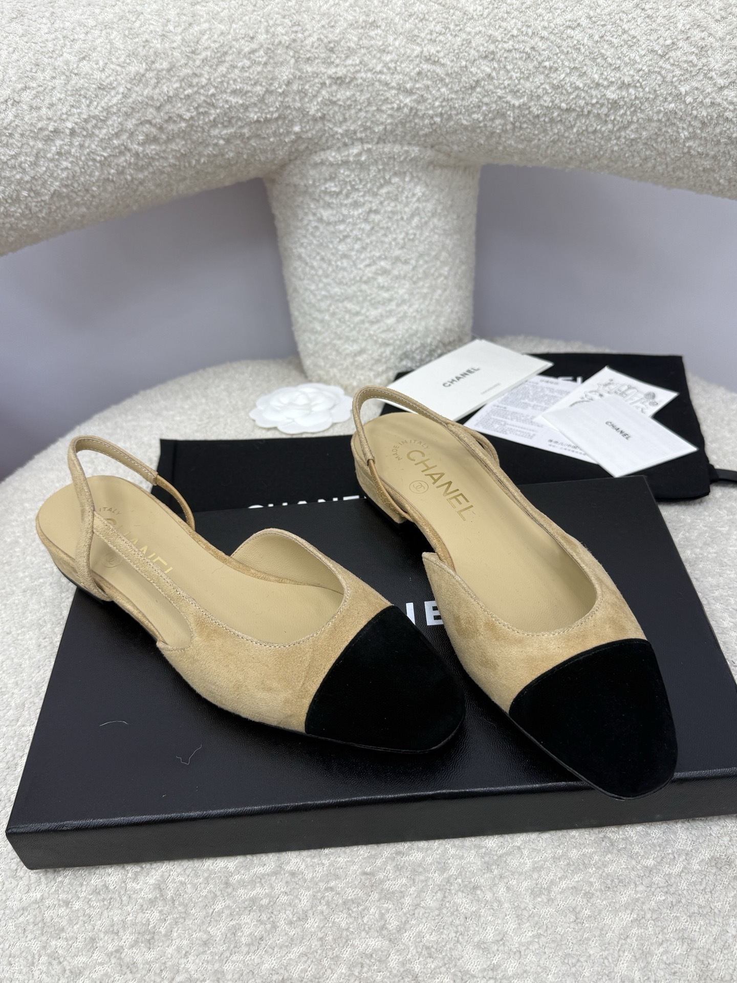 A pair of Chanel two-tone beige and black suede slingback flats displayed on a white textured surface with branded dust bags and authentication cards. - Hình 5