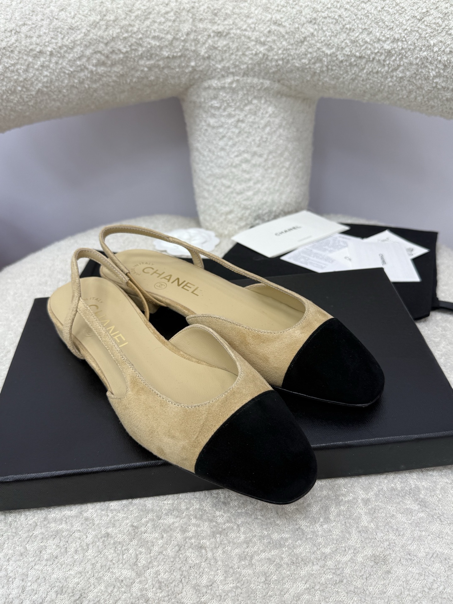 A pair of Chanel two-tone beige and black suede slingback flats displayed on a white textured surface with branded dust bags and authentication cards. - Hình 4