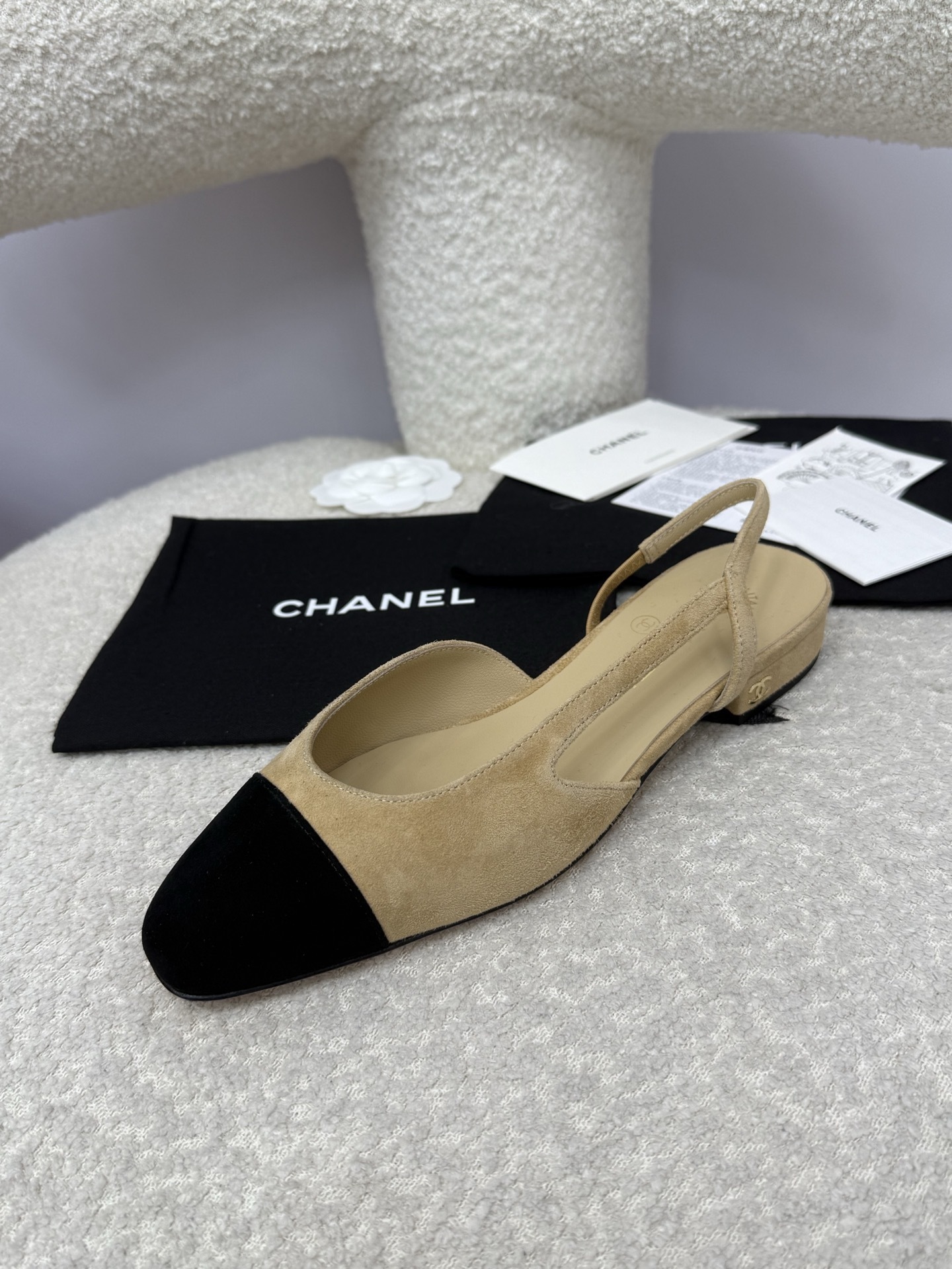 A pair of Chanel two-tone beige and black suede slingback flats displayed on a white textured surface with branded dust bags and authentication cards. - Hình 3