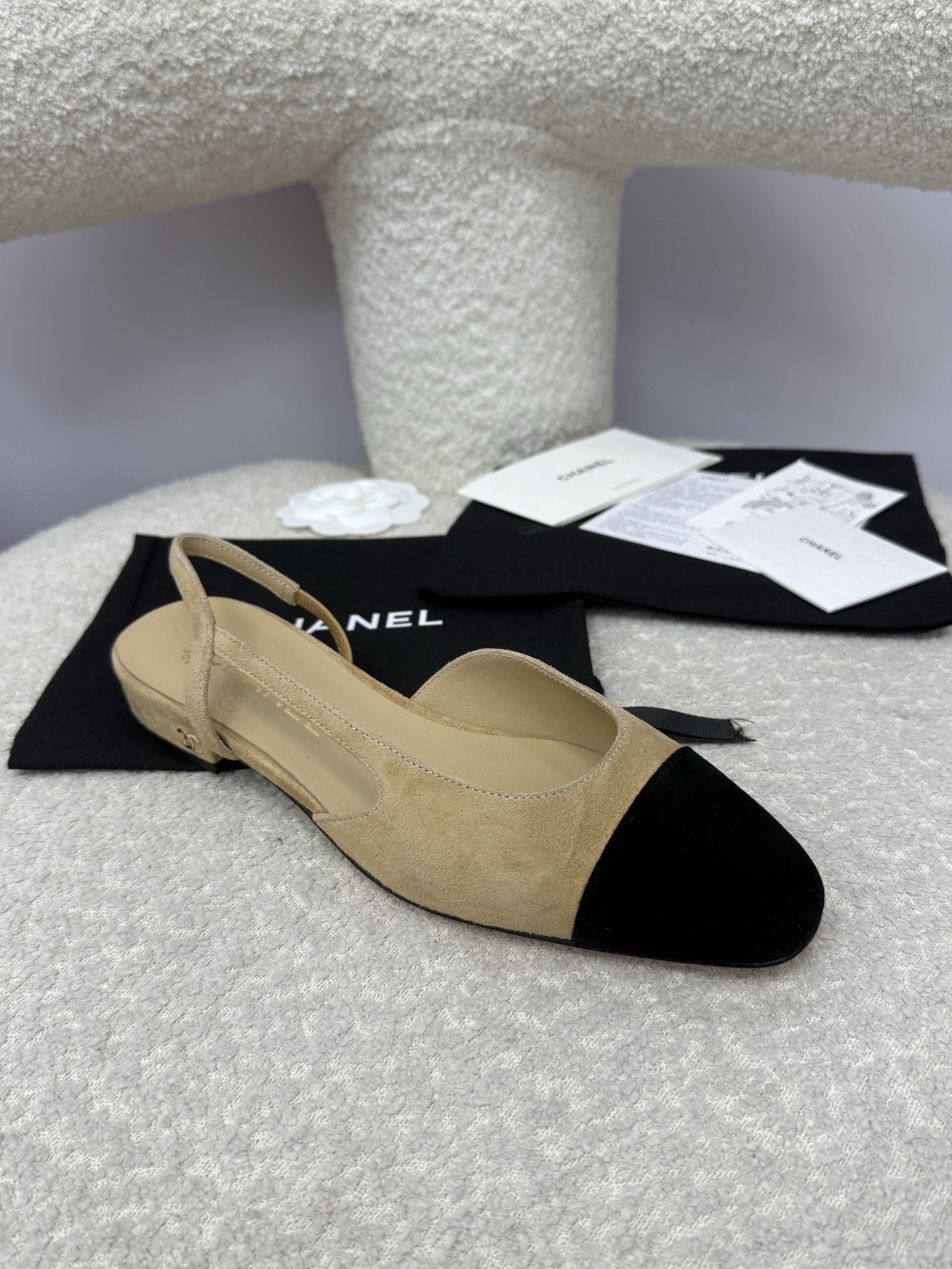 A pair of Chanel two-tone beige and black suede slingback flats displayed on a white textured surface with branded dust bags and authentication cards. - Hình 2