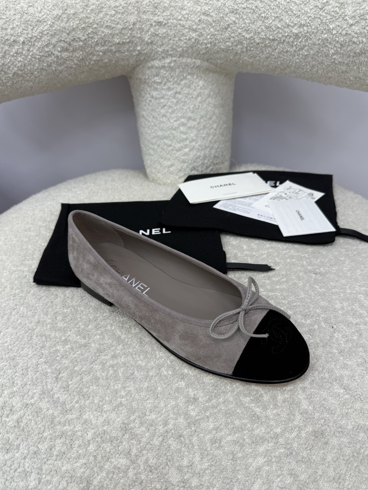 Pair of Chanel grey suede ballerina flats with black suede cap-toe and bow detail on a white textured surface. - Hình 2