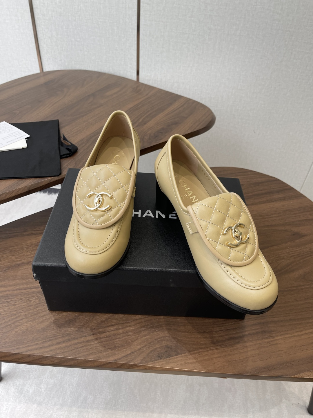 A pair of Chanel beige leather loafers with quilted gold CC logo displayed on a black shoe box. - Hình 4