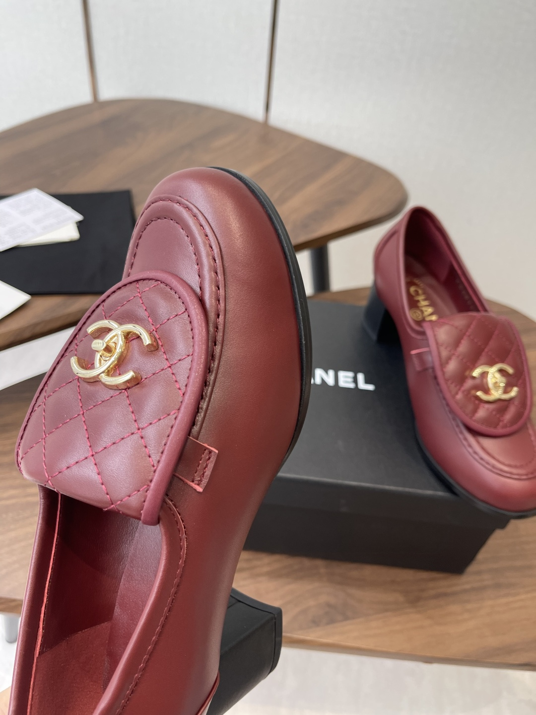 A pair of luxury burgundy Chanel quilted leather loafers with gold CC logos displayed on a black shoe box. - Hình 6