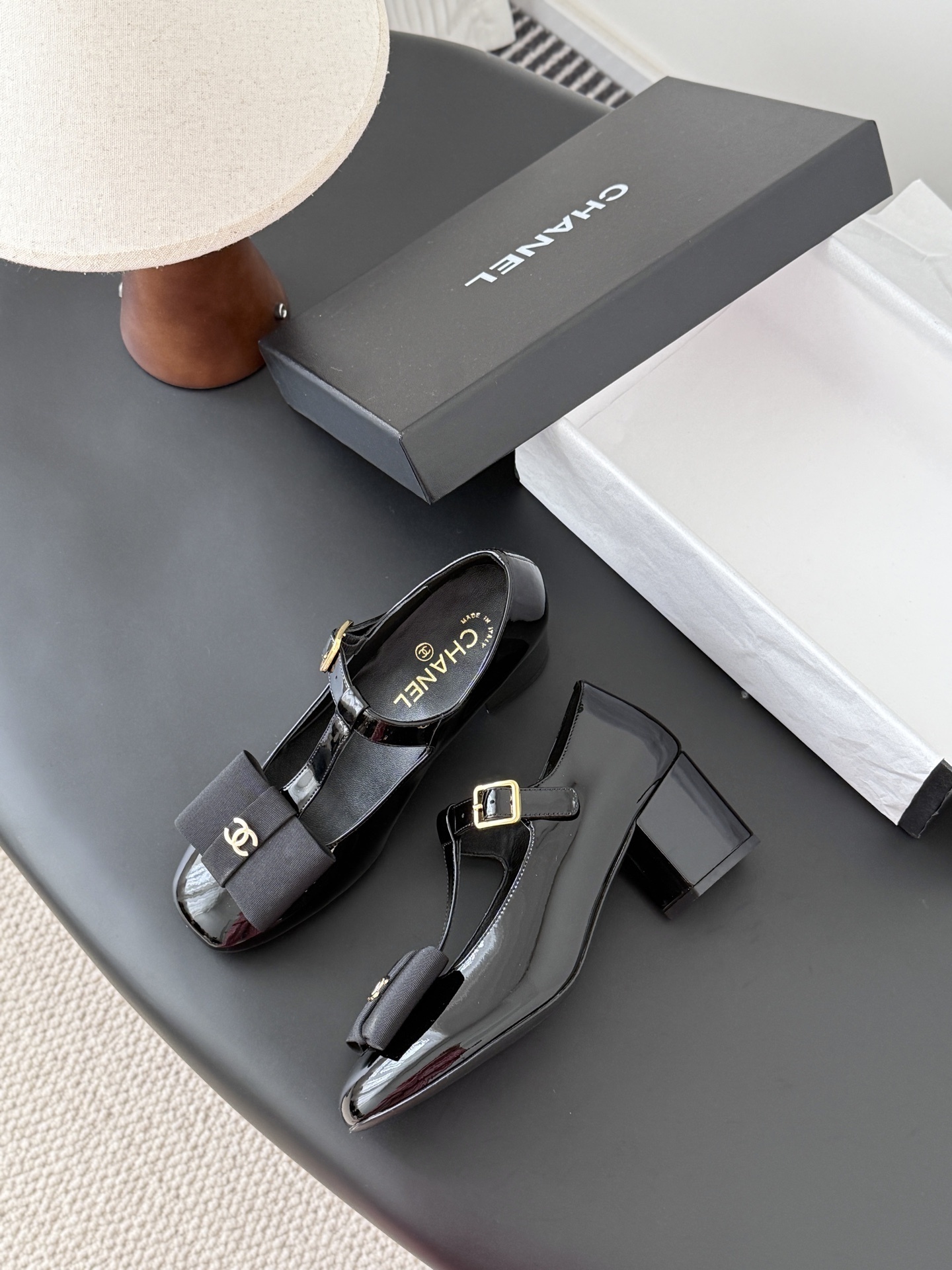 Pair of black Chanel patent leather Mary Jane pumps with T-strap, gold CC logo bow, and block heel next to a Chanel shoe box. - Hình 4