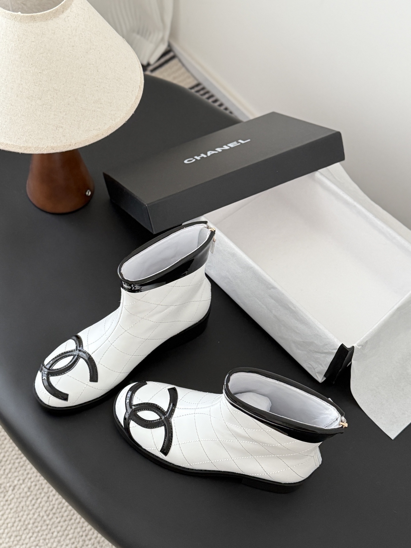 Pair of Chanel white quilted leather ankle boots with black patent leather toe caps and interlocking CC logos on a black surface. - Hình 6