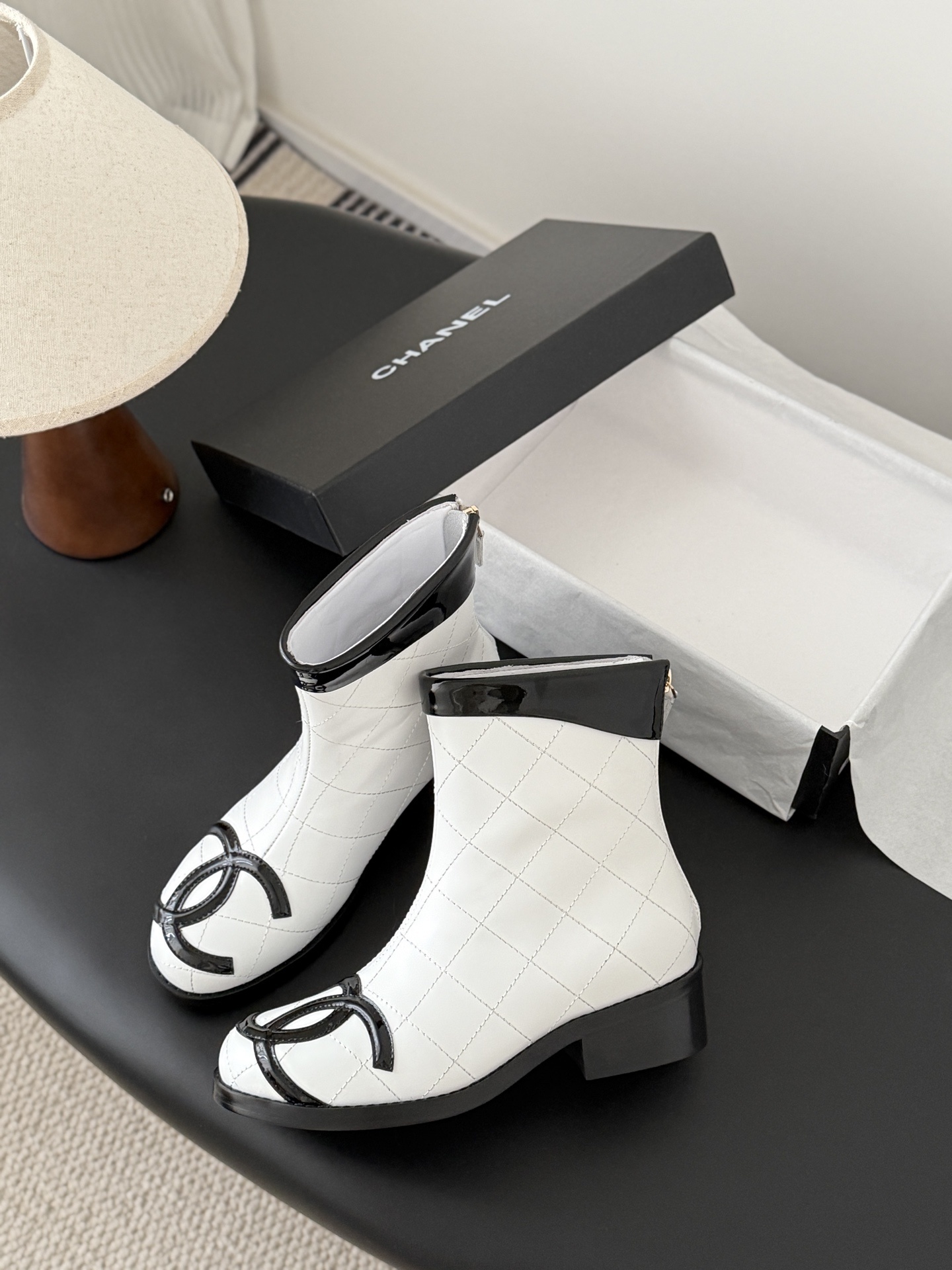 Pair of Chanel white quilted leather ankle boots with black patent leather toe caps and interlocking CC logos on a black surface. - Hình 4