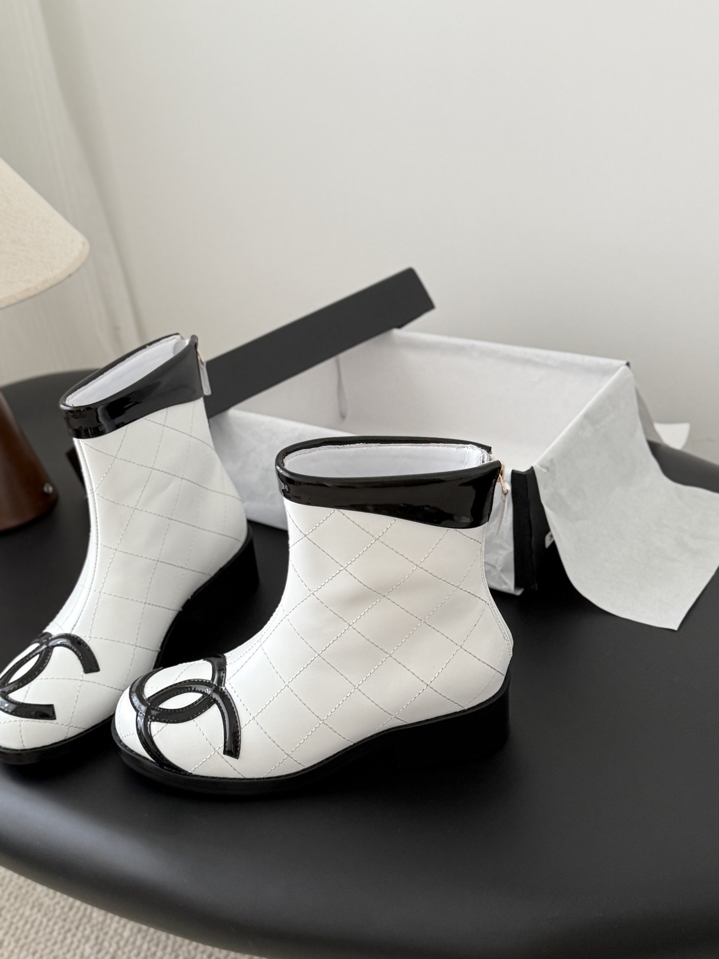 Pair of Chanel white quilted leather ankle boots with black patent leather toe caps and interlocking CC logos on a black surface. - Hình 2