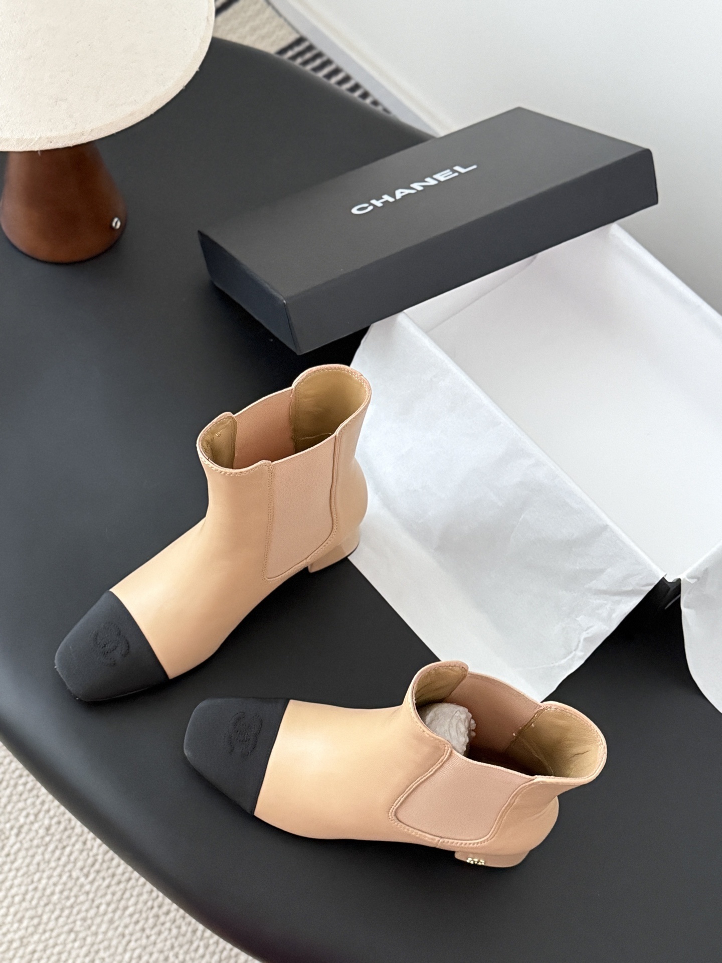 Chanel beige leather ankle boots with black toe caps and block heels displayed next to a black Chanel shoe box. - Hình 5