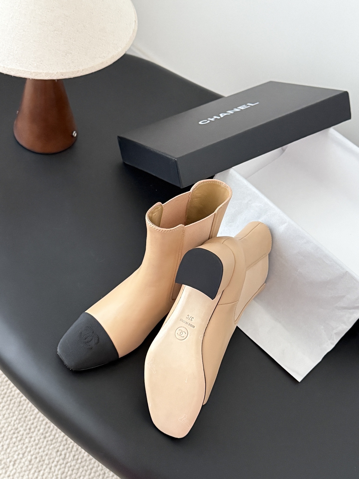 Chanel beige leather ankle boots with black toe caps and block heels displayed next to a black Chanel shoe box. - Hình 4