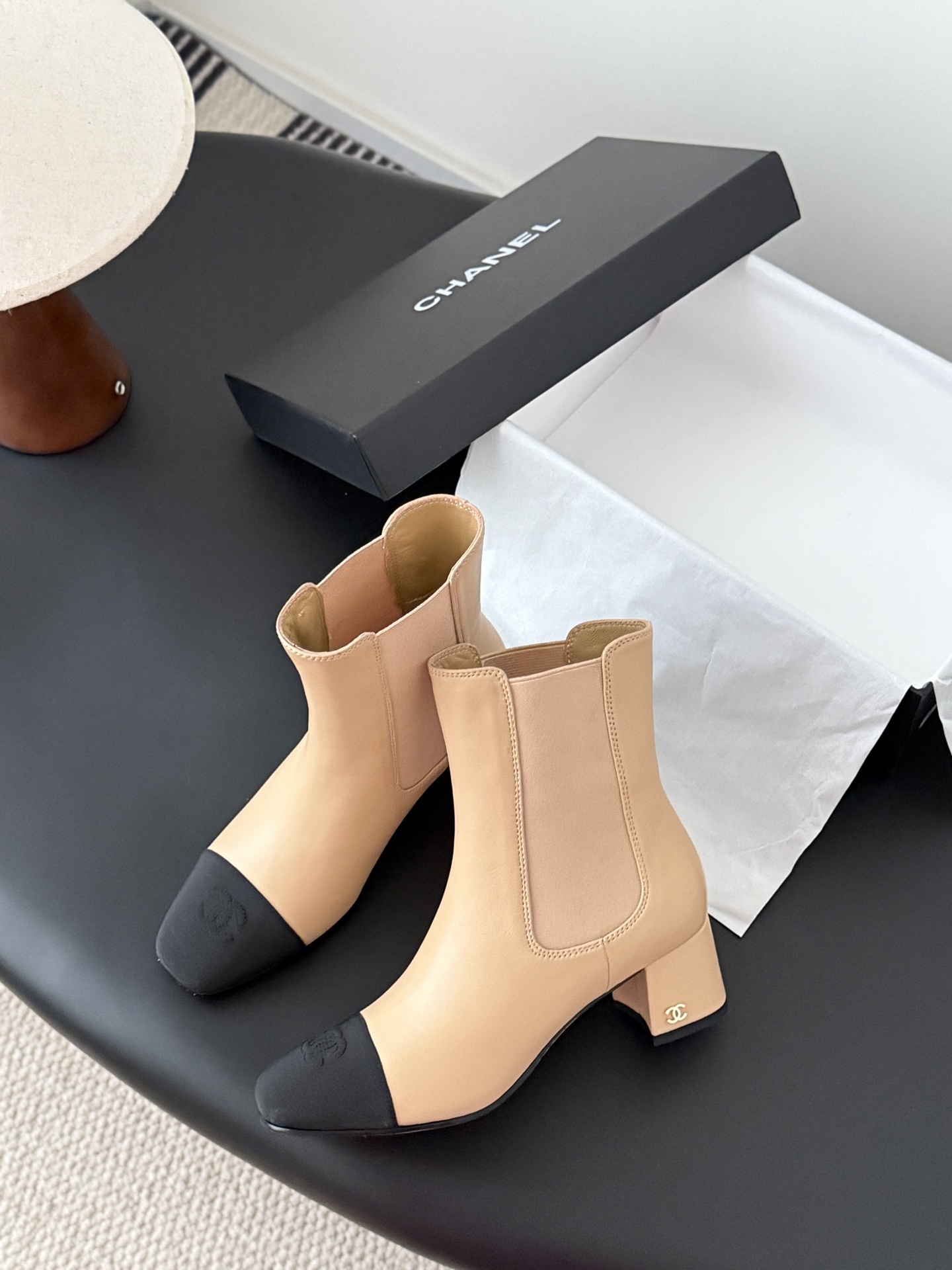 Chanel beige leather ankle boots with black toe caps and block heels displayed next to a black Chanel shoe box. - Hình 3
